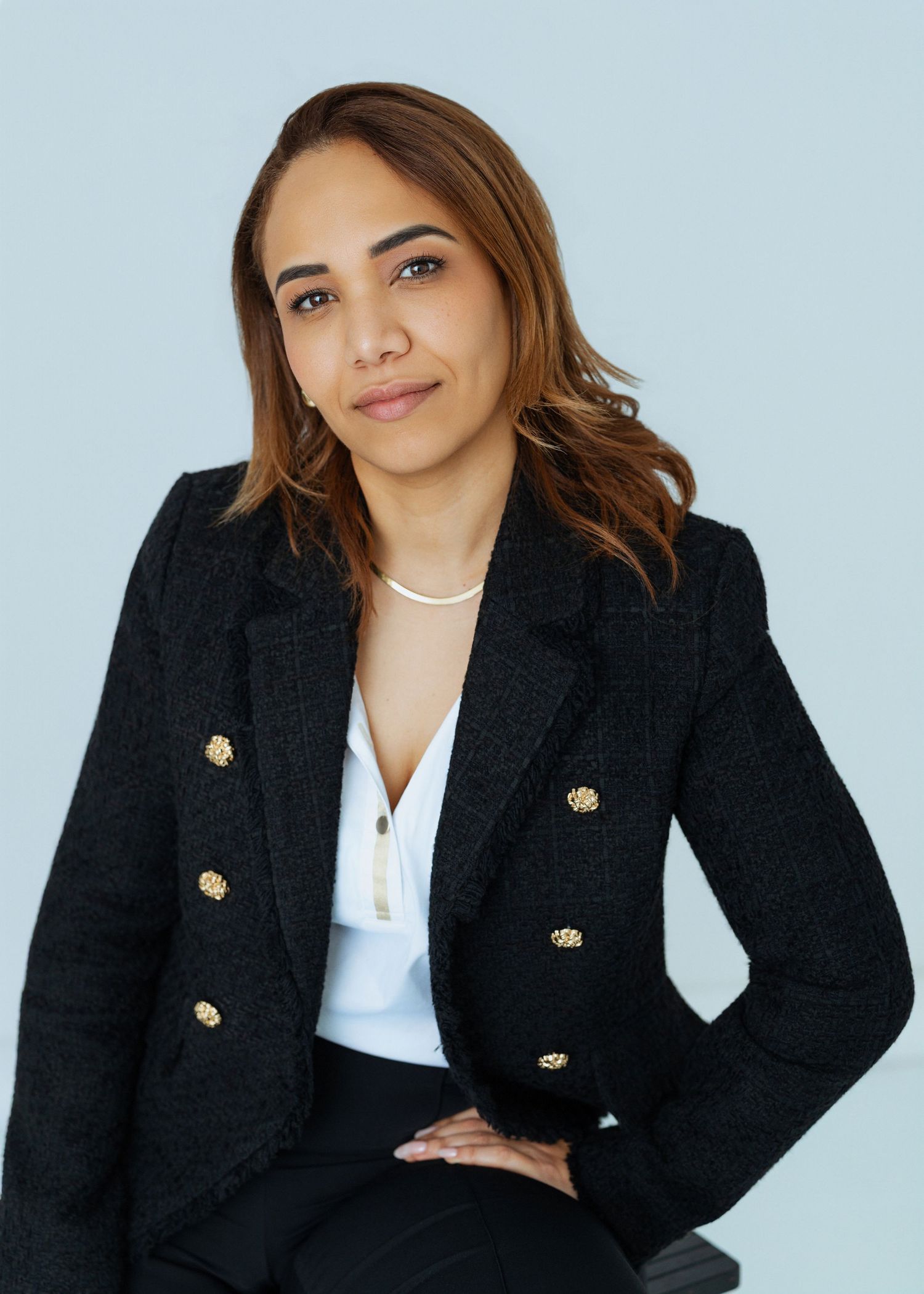 Professional wearing a classic black blazer with gold buttons and white top poses against light background.