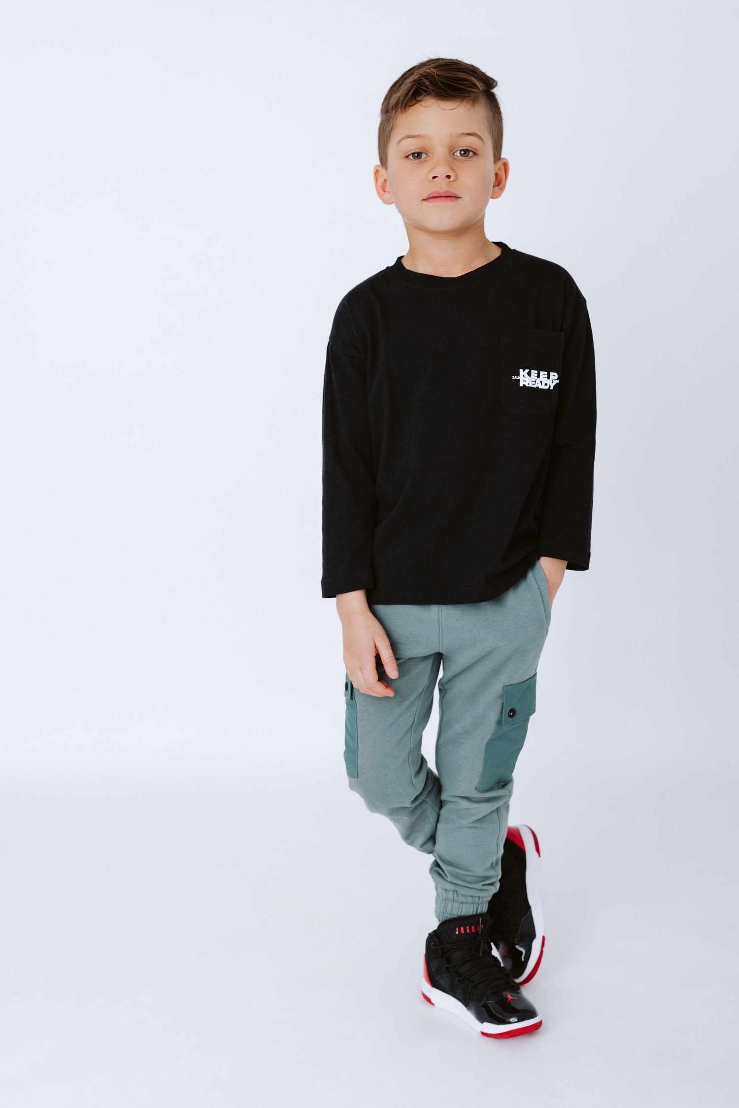 Young boy in black long sleeve shirt and cargo pants posing against white background.