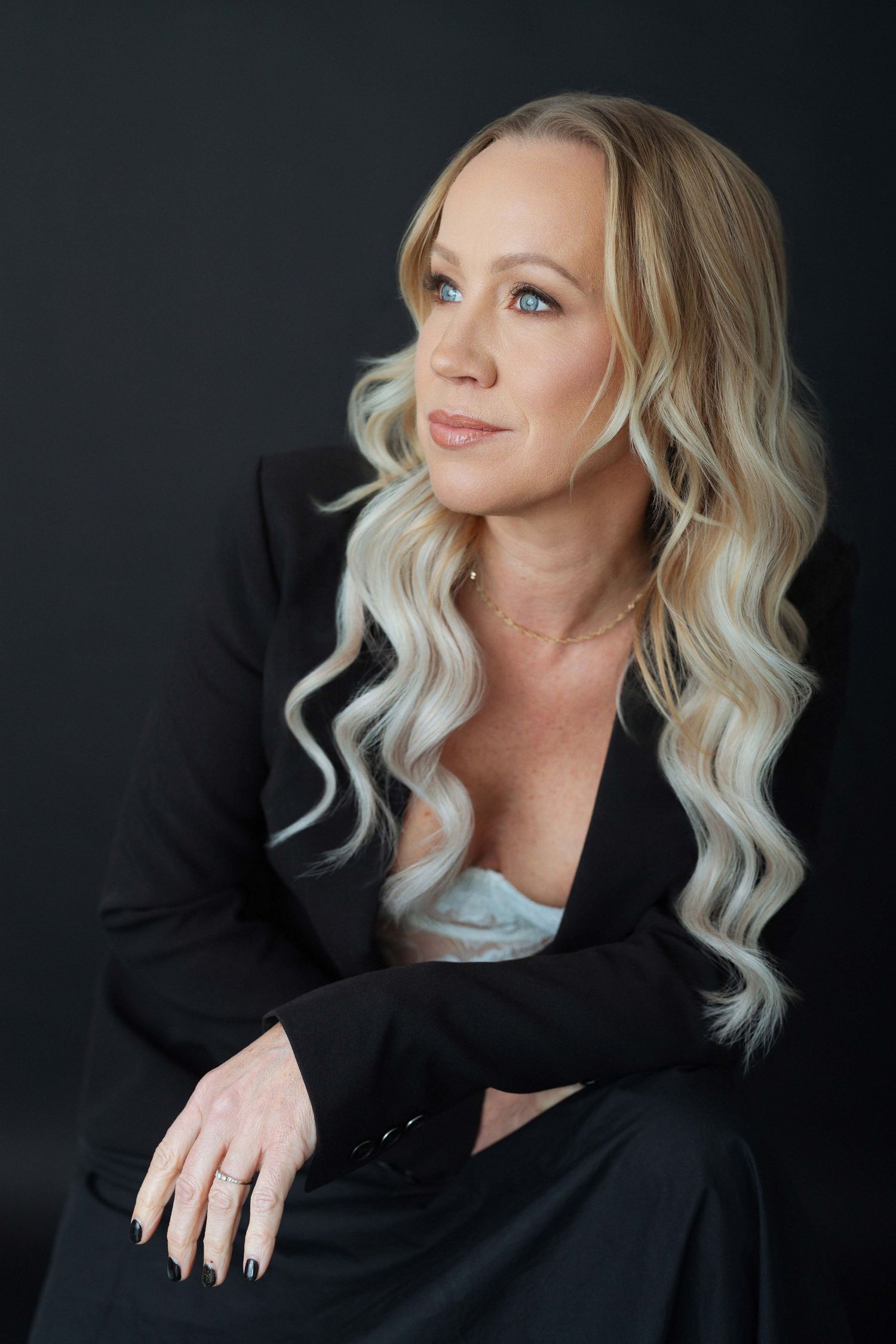 Blonde woman in black blazer with wavy hair poses confidently against dark background in professional portrait.
