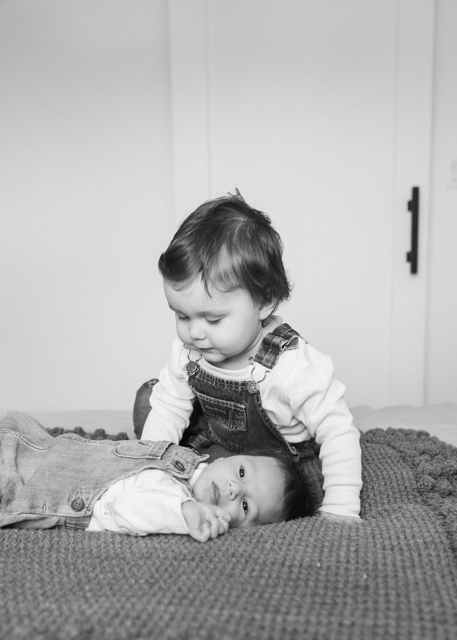 Black and white photo of a toddler in overalls sitting beside a newborn baby lying on a textured blanket.