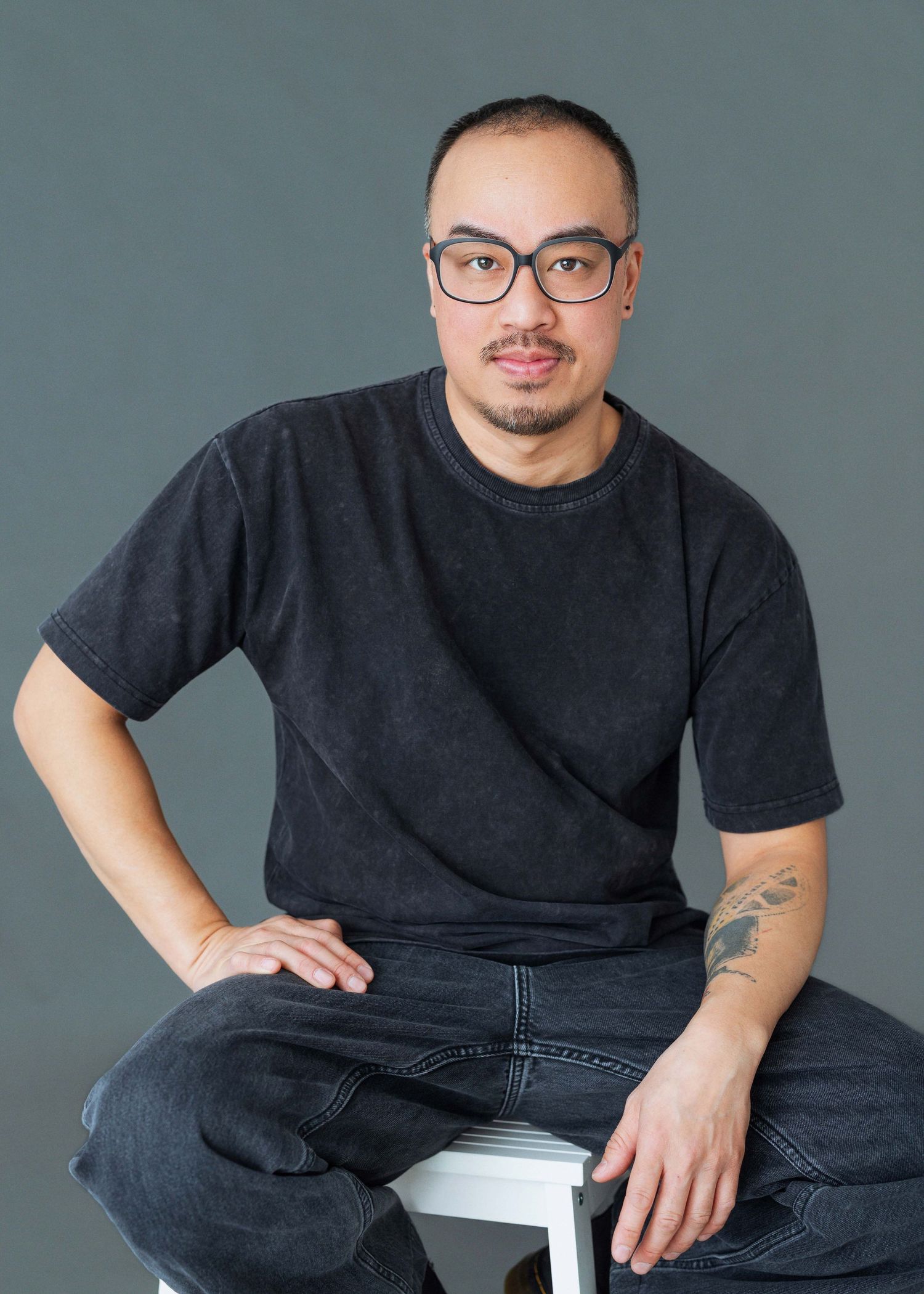 Person in black t-shirt and jeans sits casually on white chair against gray backdrop in professional photo series.