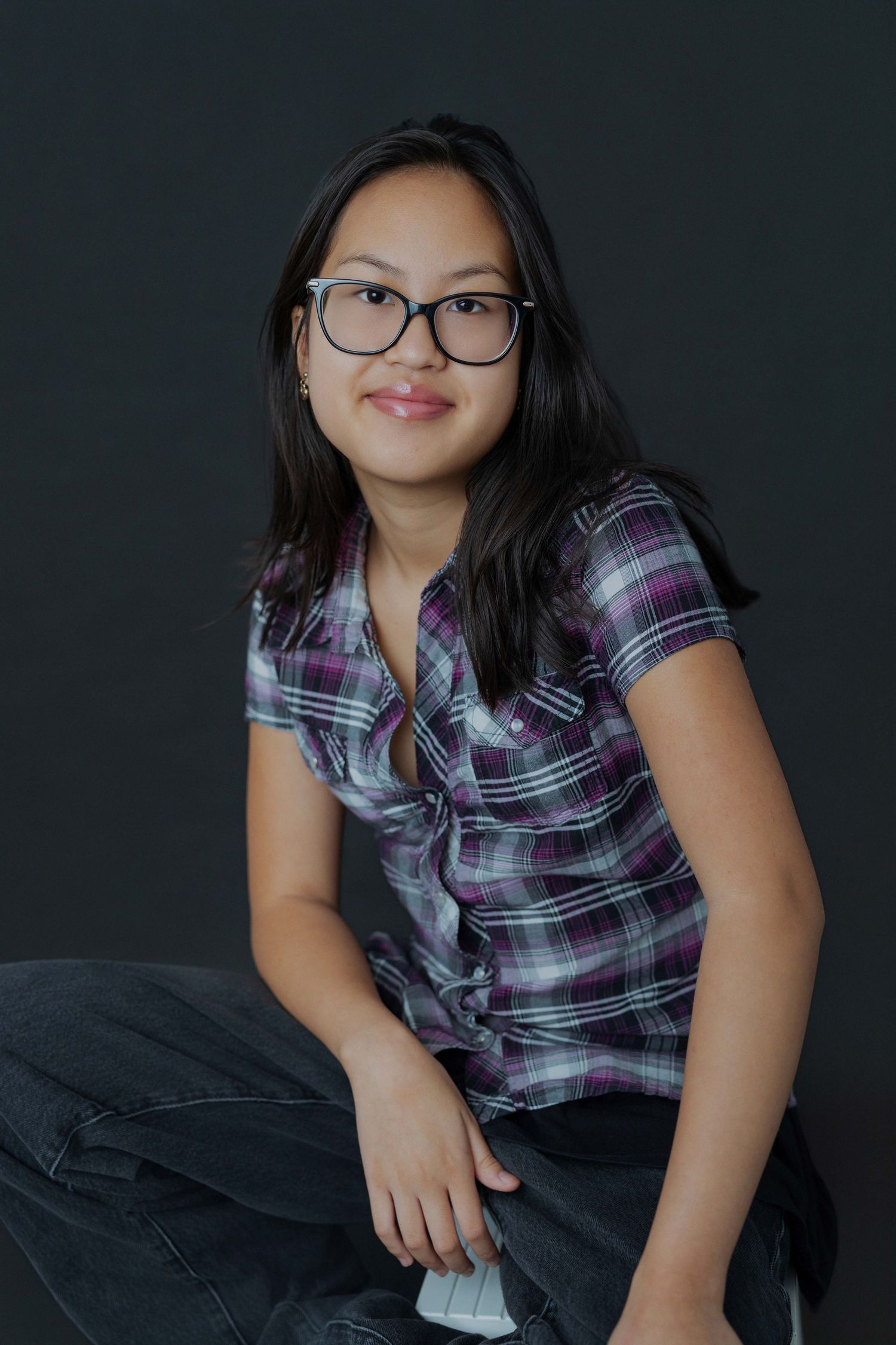 Young woman with glasses and plaid shirt sitting confidently against dark background.