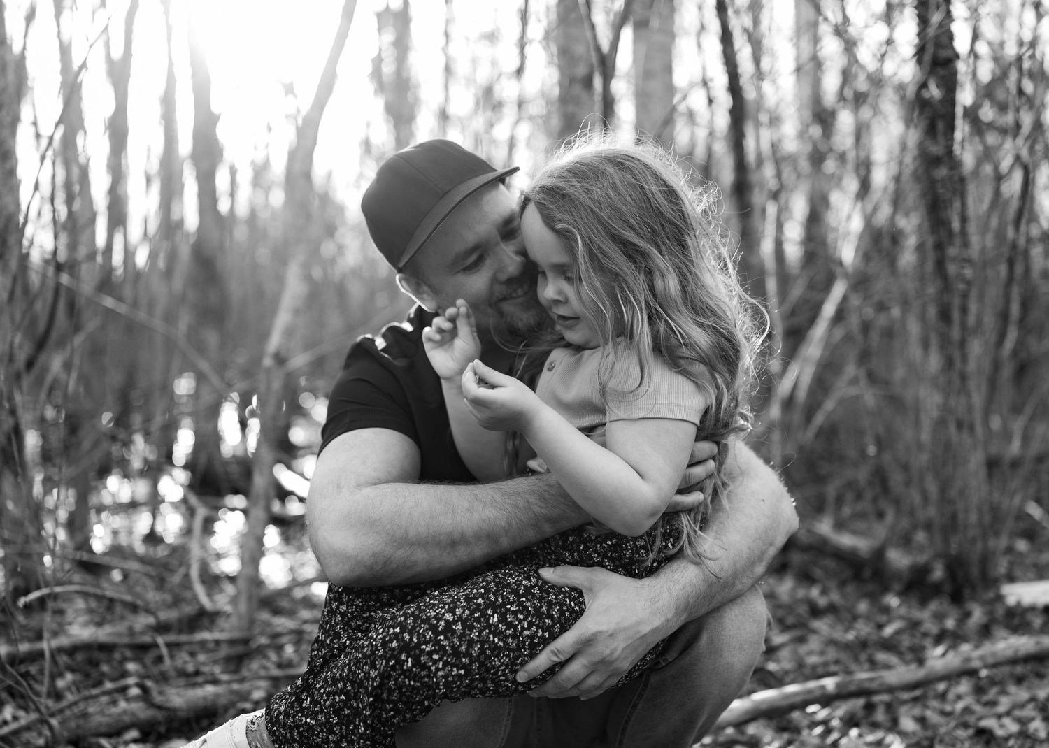 Black and white photo of a father tenderly embracing his young daughter outdoors in a wooded forest setting.