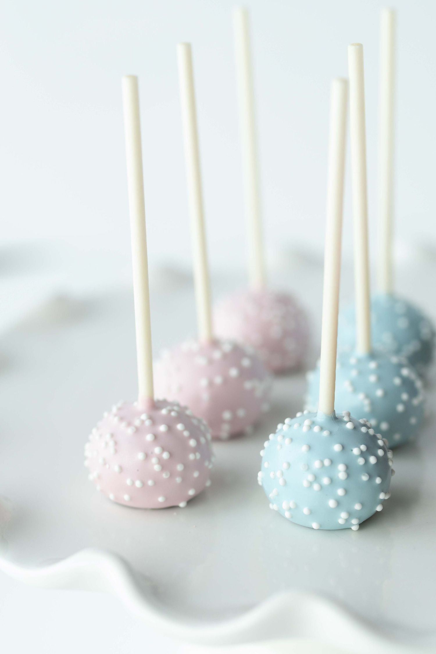 Pink and blue cake pops decorated with white pearl sprinkles on wooden sticks against a light background.