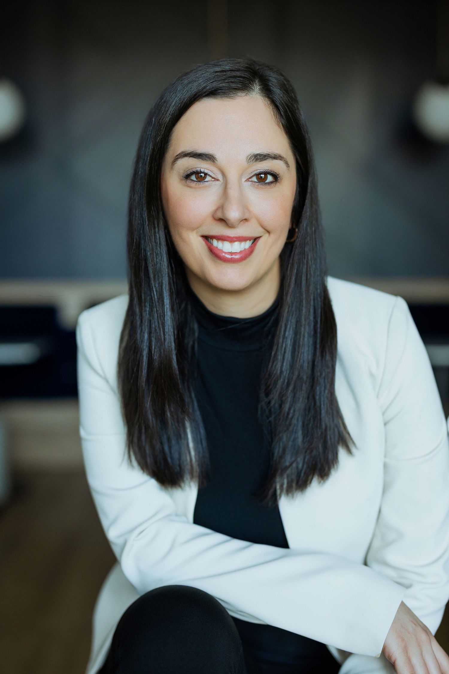 Professional woman in white blazer and black top smiling confidently with arms crossed in modern office setting.