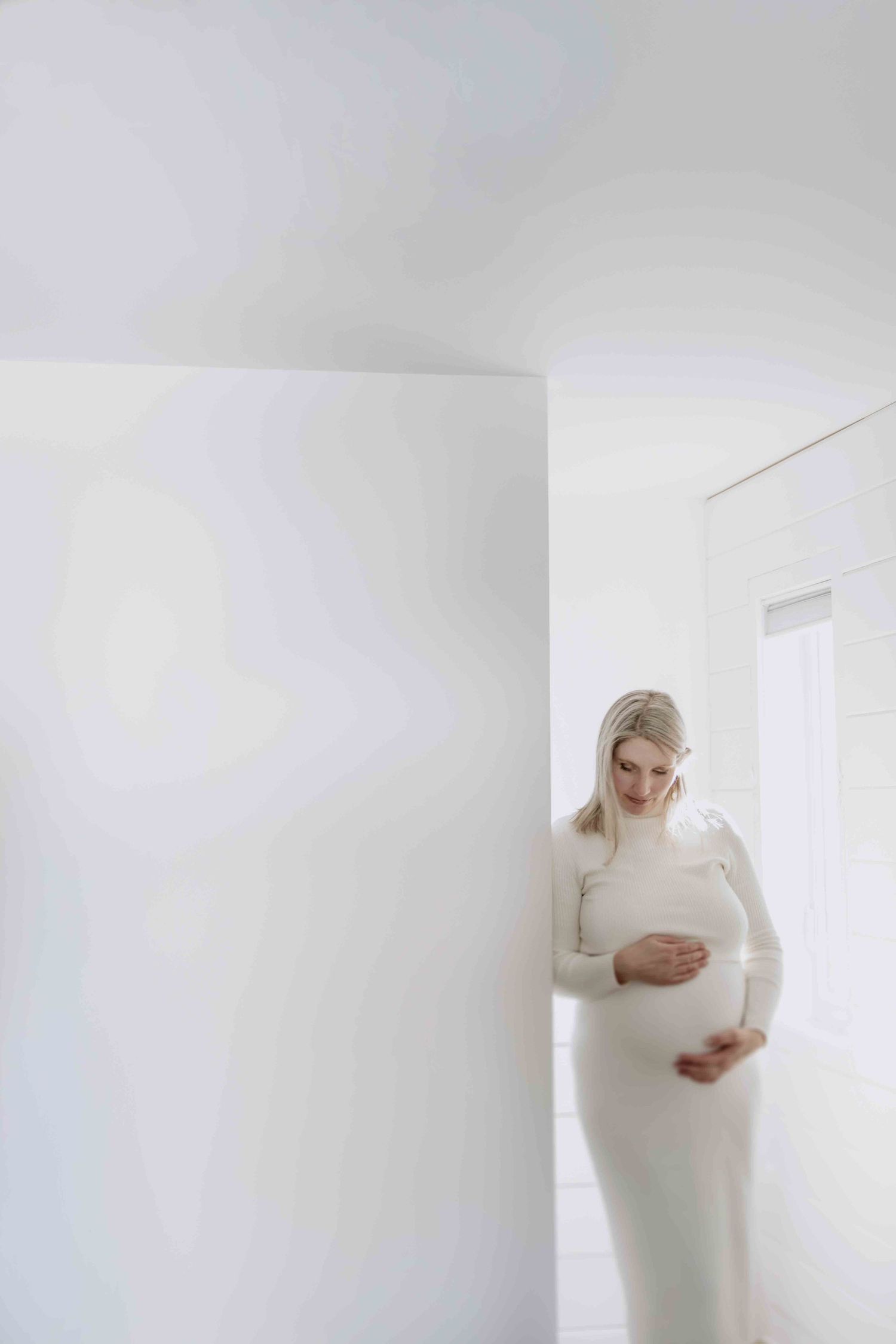Expectant mother in white dress poses against minimalist white wall interior.