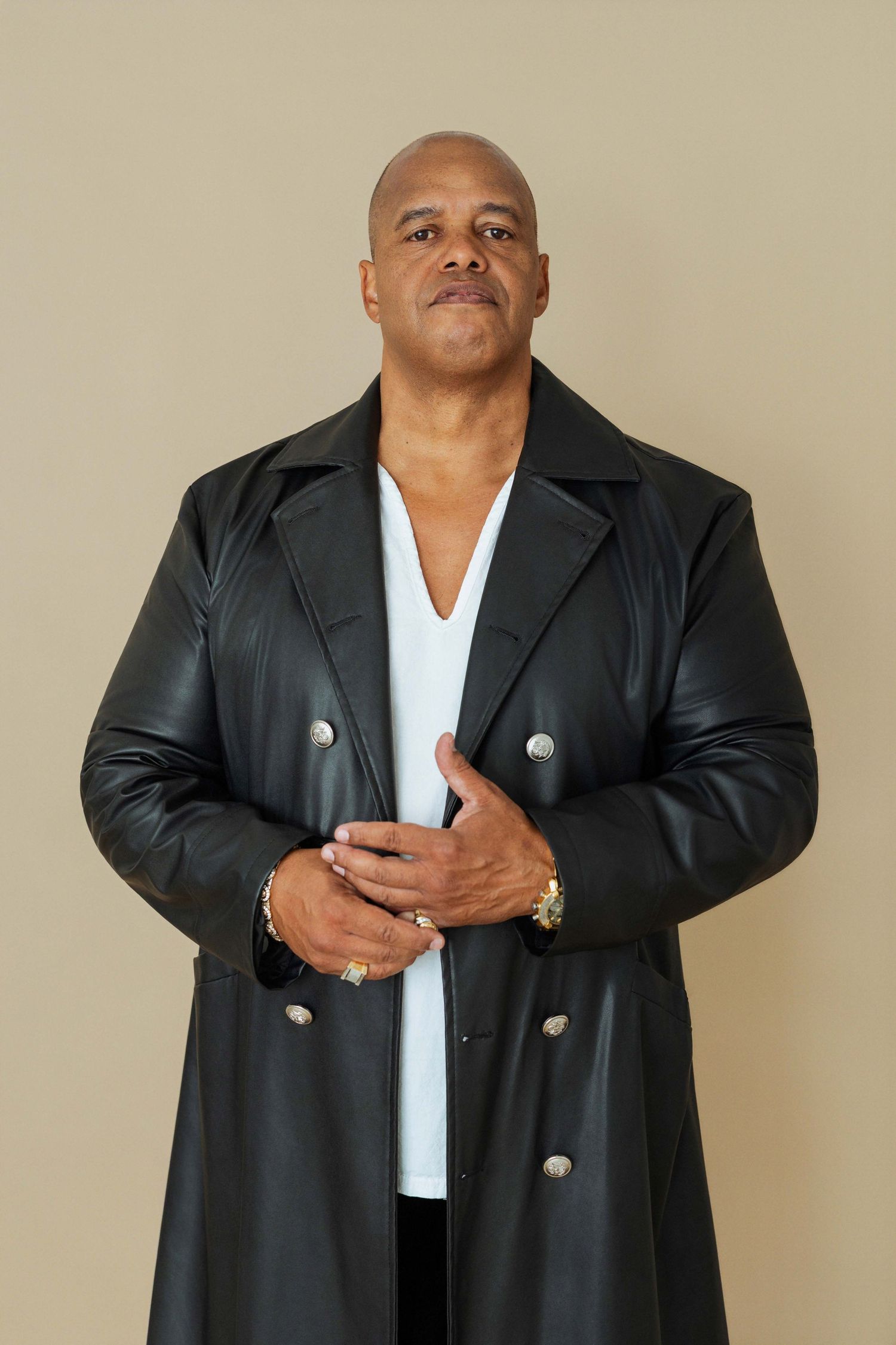 casting Bald man in black leather trench coat with silver buttons over white V-neck shirt, posing confidently against beige background.