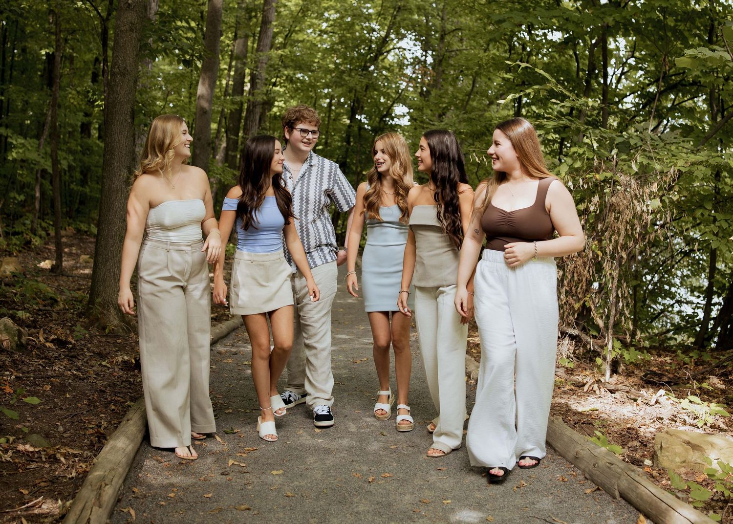 Five young adults walking together on a wooded path, smiling and enjoying a casual outdoor stroll.
