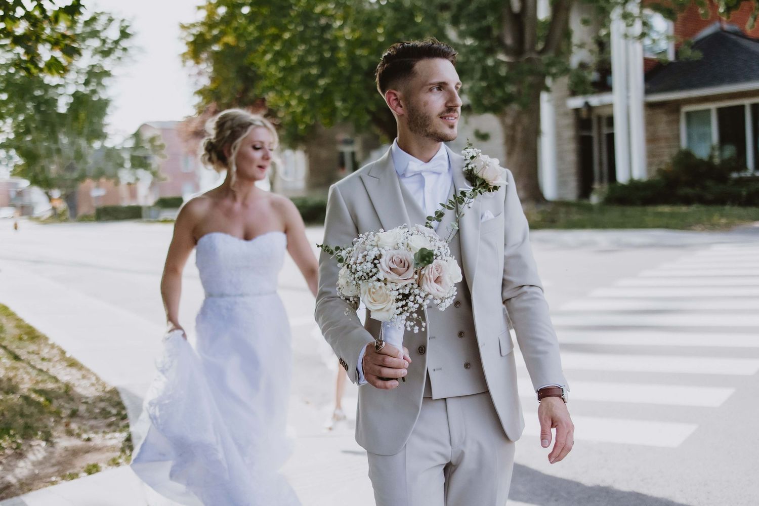 Candid wedding photo of a couple walking, with the groom in light gray suit carrying white flowers.