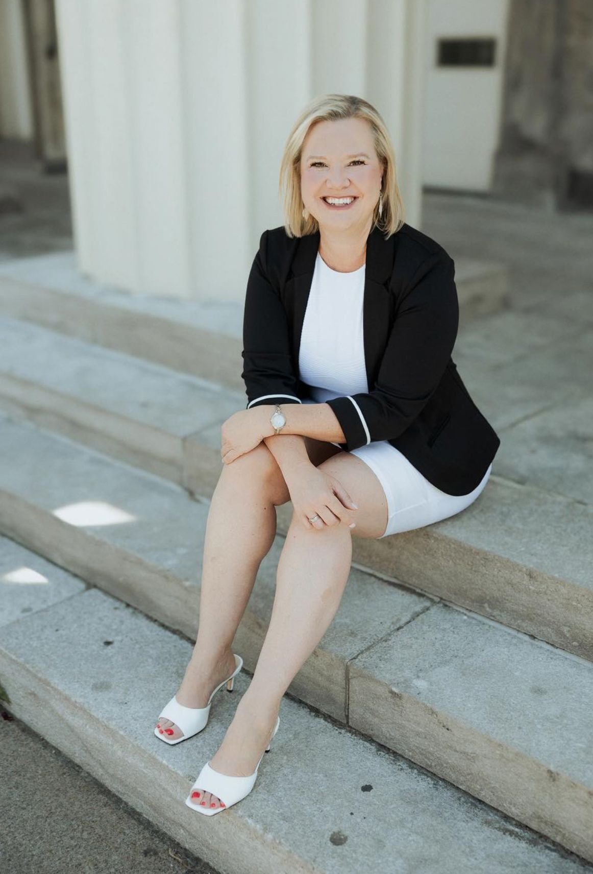 Professional lifestyle photo showing legs in white sandals and black blazer sitting on concrete steps.