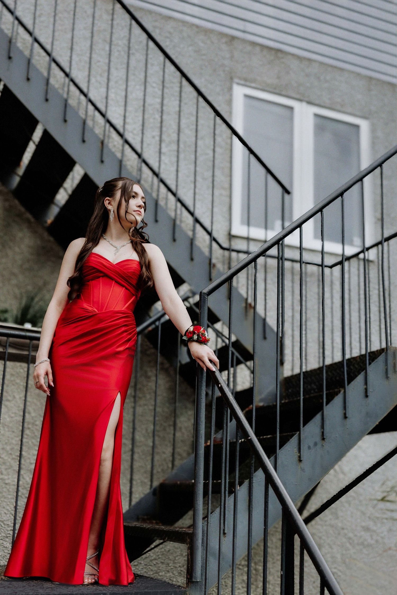 Young woman in elegant red satin gown with slit poses on outdoor metal staircase, wearing floral wrist corsage.