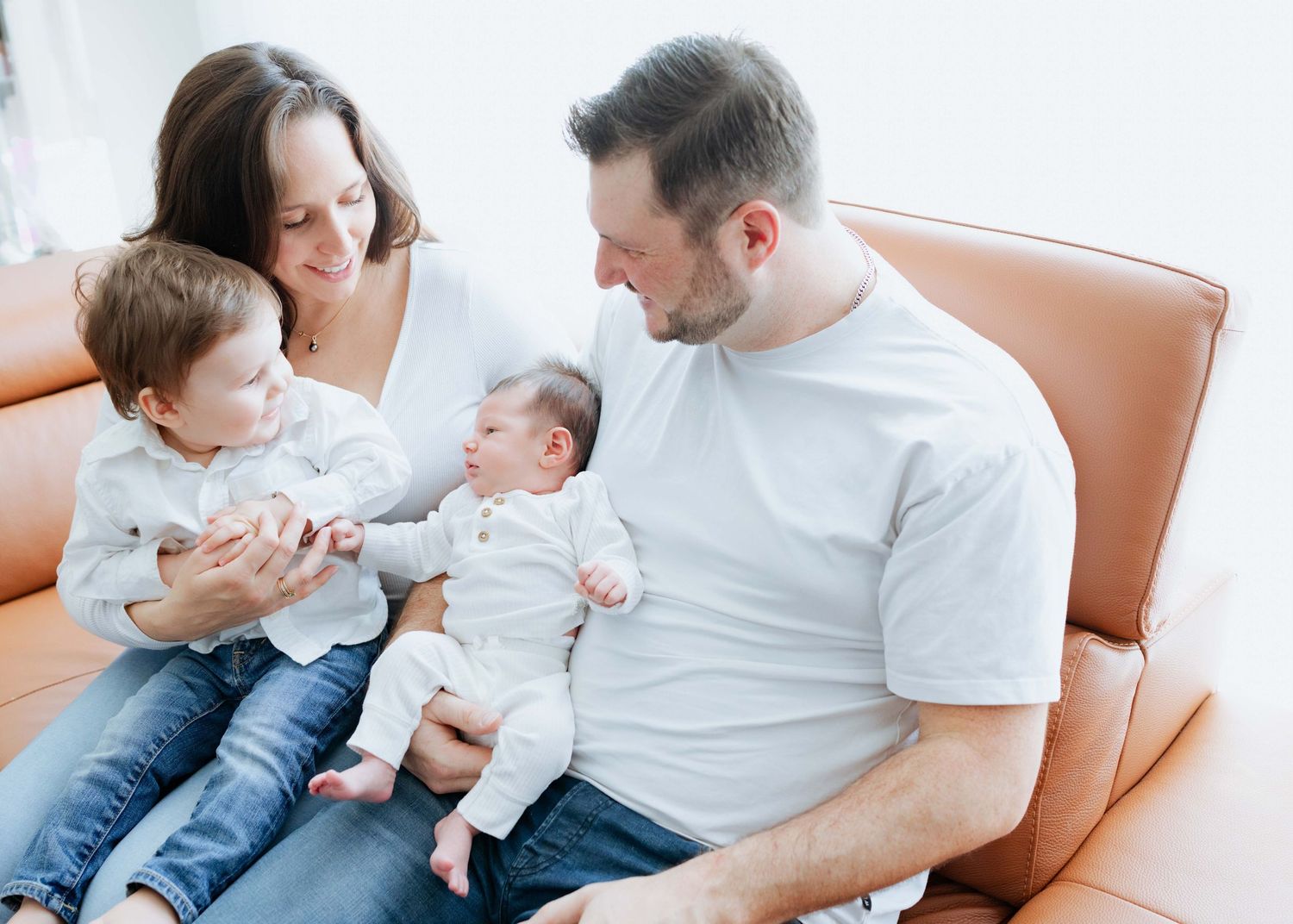 Happy family with two parents sitting on a brown leather sofa holding a toddler and newborn baby.