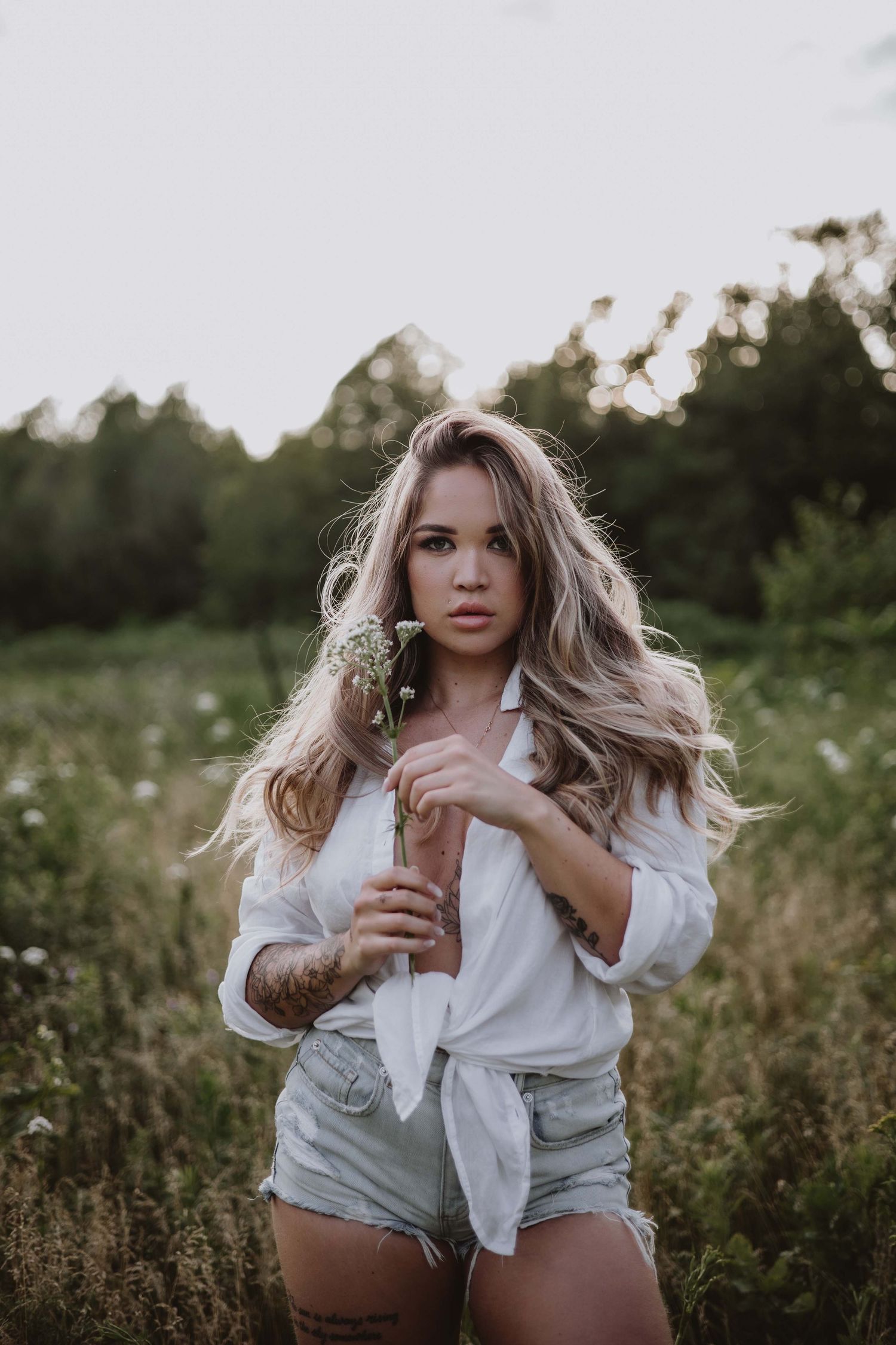 Woman in white shirt and denim shorts poses in wildflower field during sunset.