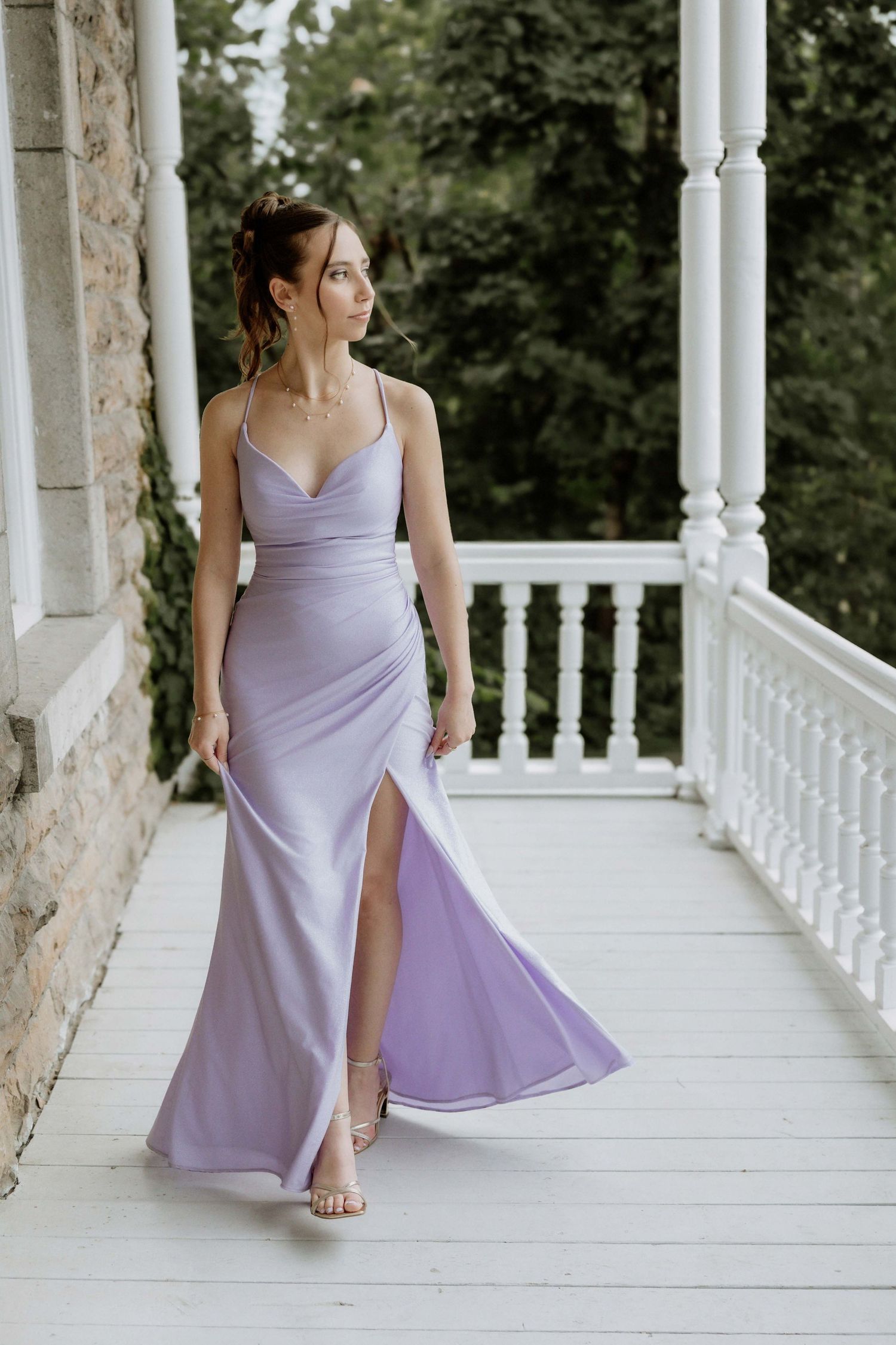 Woman in lavender satin maxi dress with slit posing on white porch balcony surrounded by lush greenery.