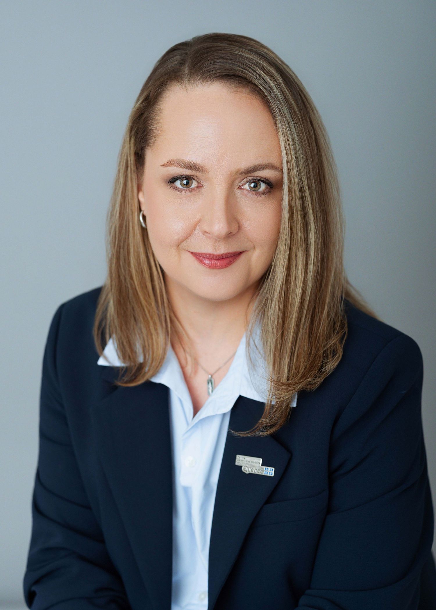 Professional headshot of a woman in a navy blazer with blonde hair, smiling confidently against a gray background.
