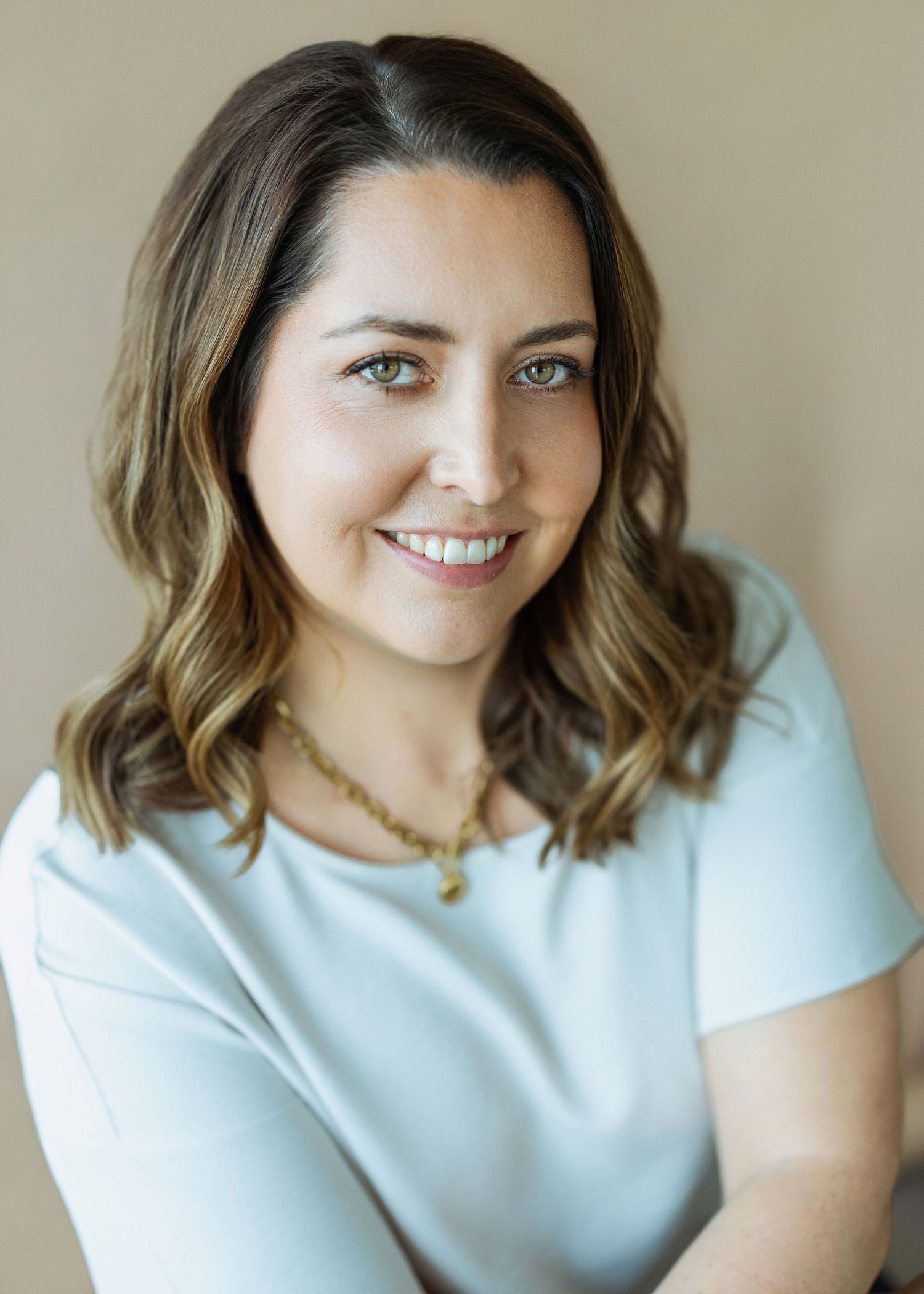 Professional headshot of a smiling woman with shoulder-length brown hair wearing a light blue top and gold necklace.