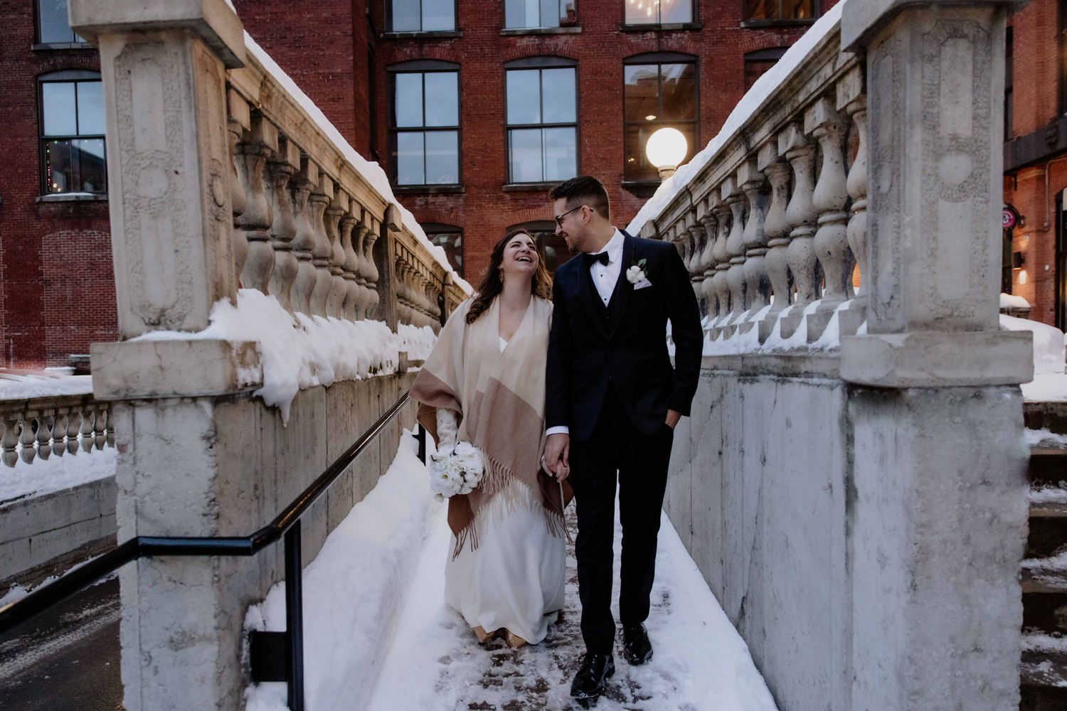 Couple in wedding attire strolling past decorative white stone railing on snowy day.
