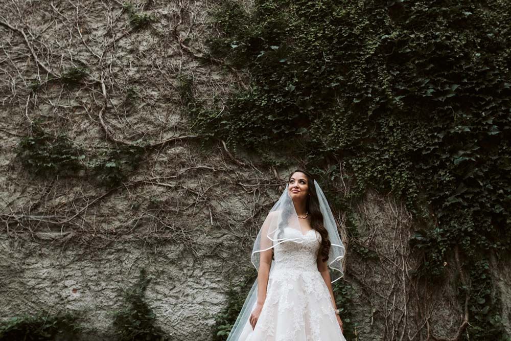 Elegant bride poses in front of stone wall wearing lace gown and flowing veil.