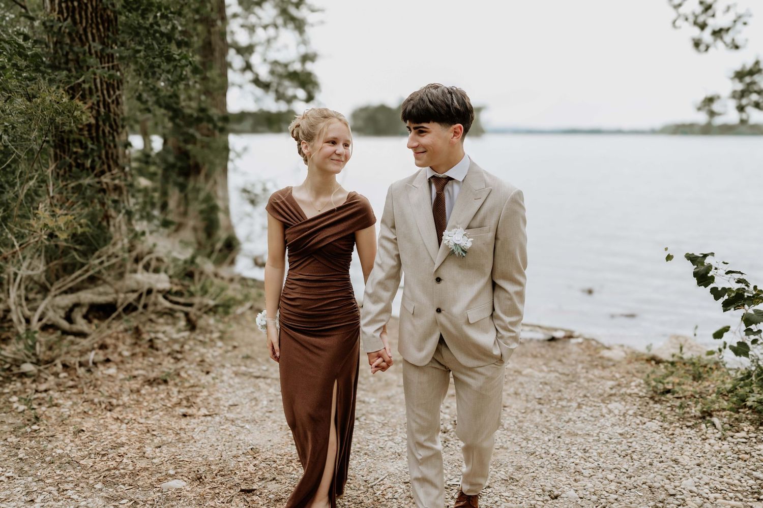 Couple walking hand in hand by a lake, woman in brown gown, man in beige suit, surrounded by lush trees.