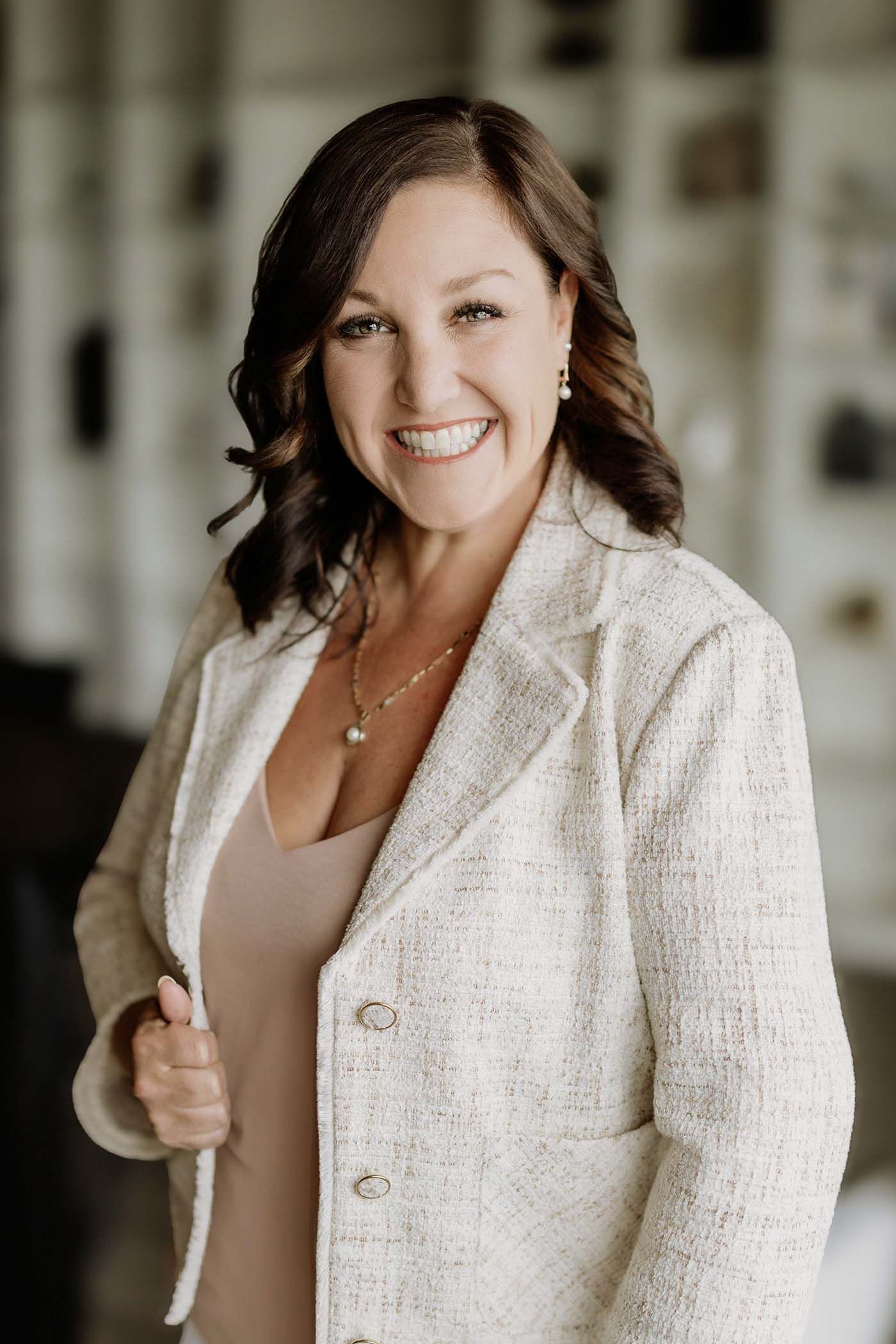 Smiling brunette woman in a cream tweed blazer poses confidently in a modern interior setting.