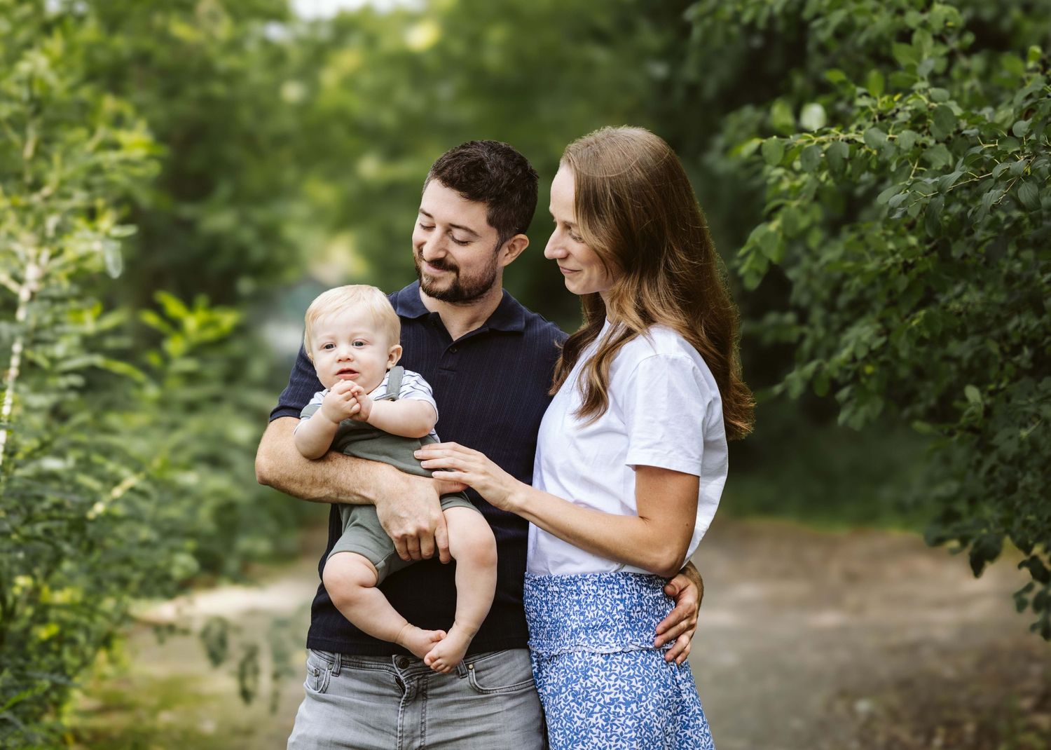 A happy young family with a baby boy poses outdoors surrounded by lush green trees on a scenic path.