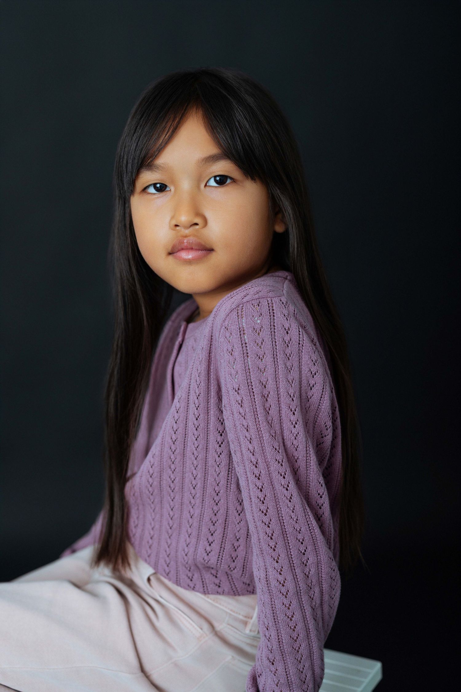 Young Asian girl with long black hair wearing a purple knit sweater, posed against a dark background.