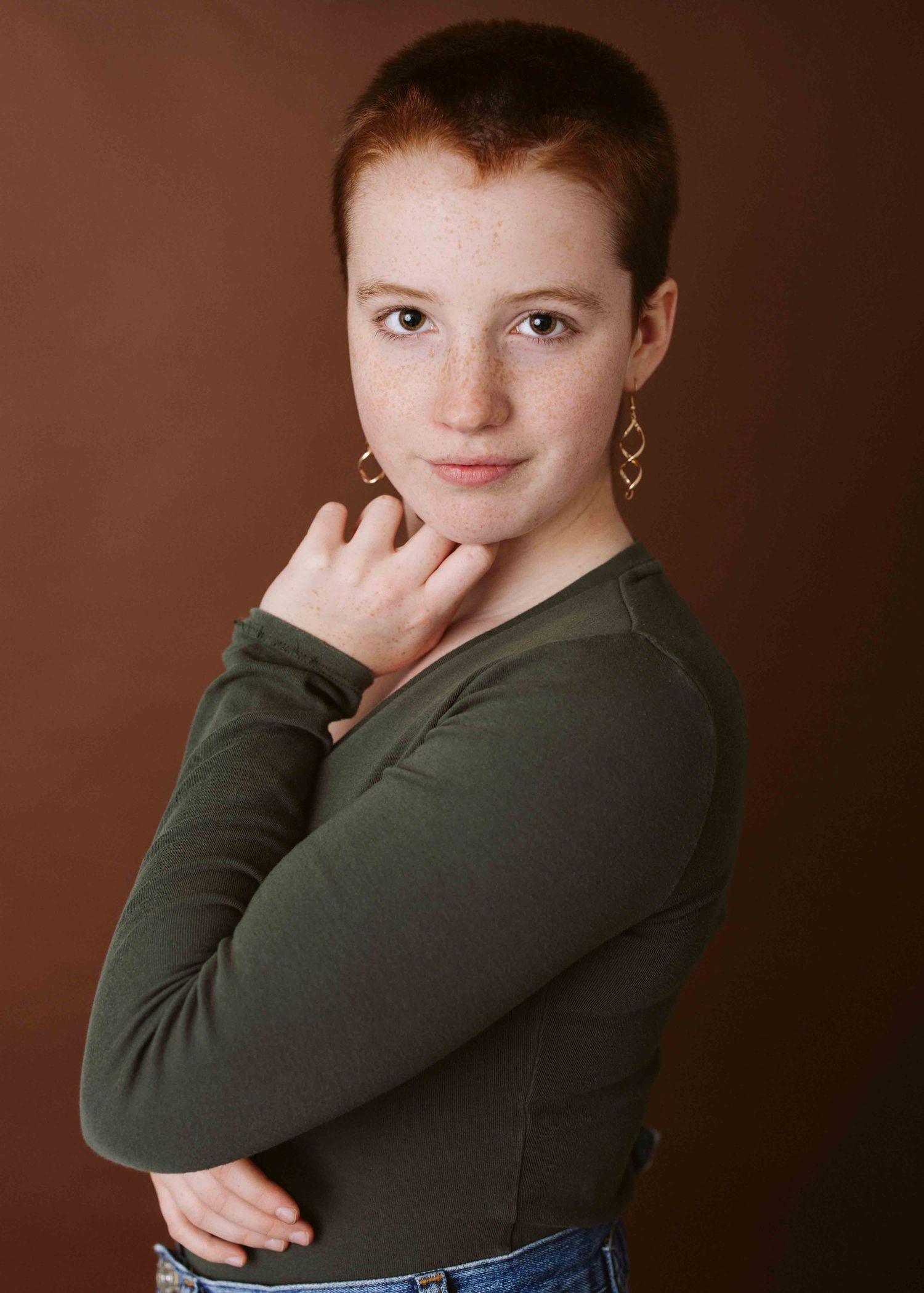 Portrait in olive green top against warm brown background with geometric earrings.