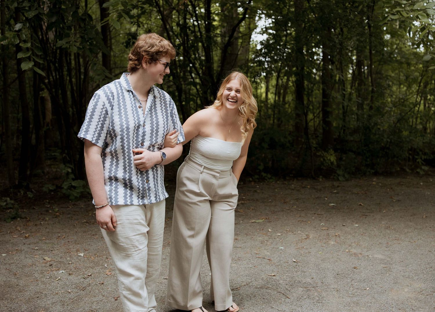 A smiling couple walks together on a path surrounded by lush green trees, dressed in casual neutral outfits.