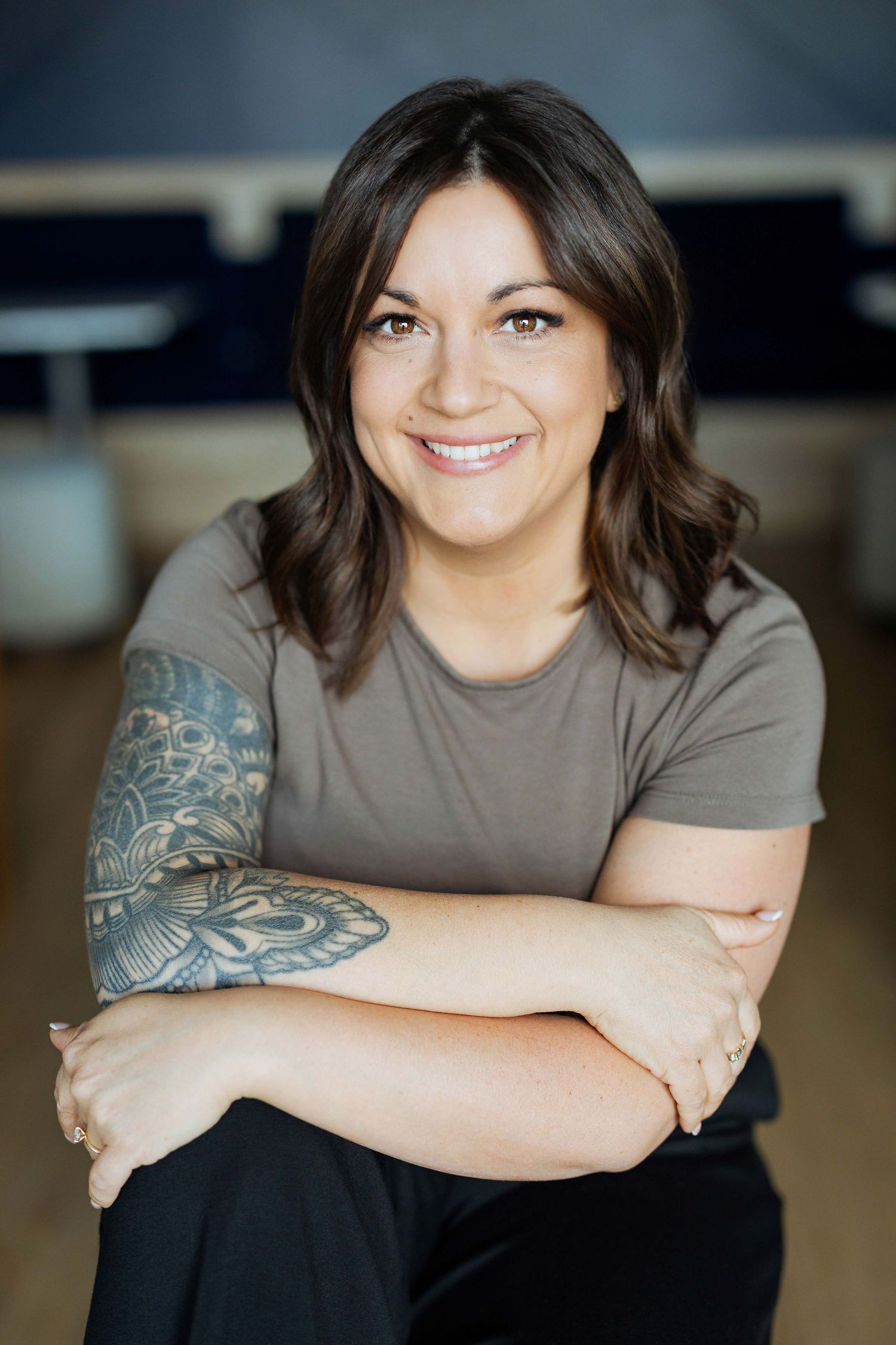 Smiling woman with dark hair and tattoo sleeve wearing gray t-shirt, sitting with arms crossed in casual indoor setting.