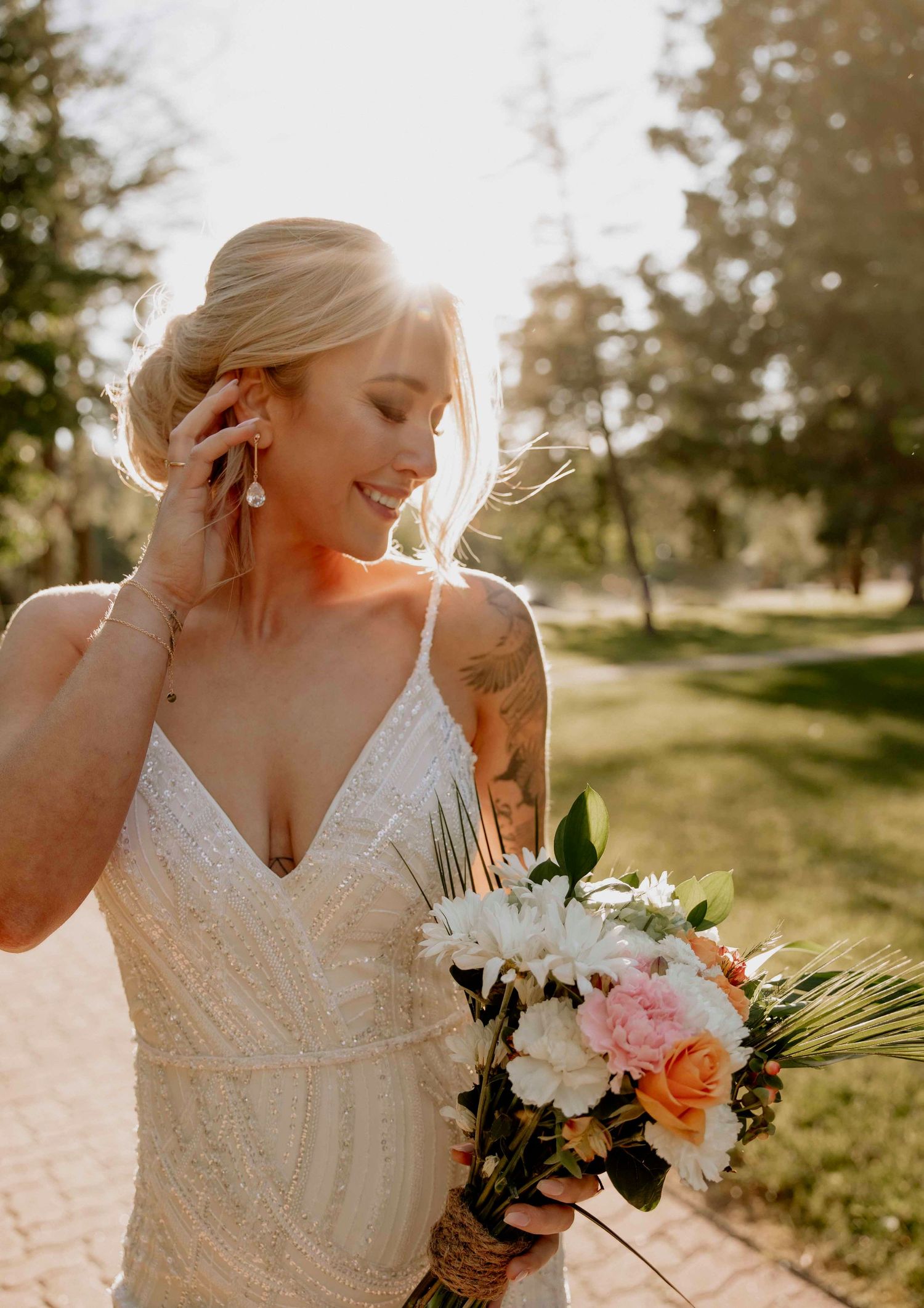 Bride in lace dress holds bouquet of pink and orange flowers in golden sunlight.