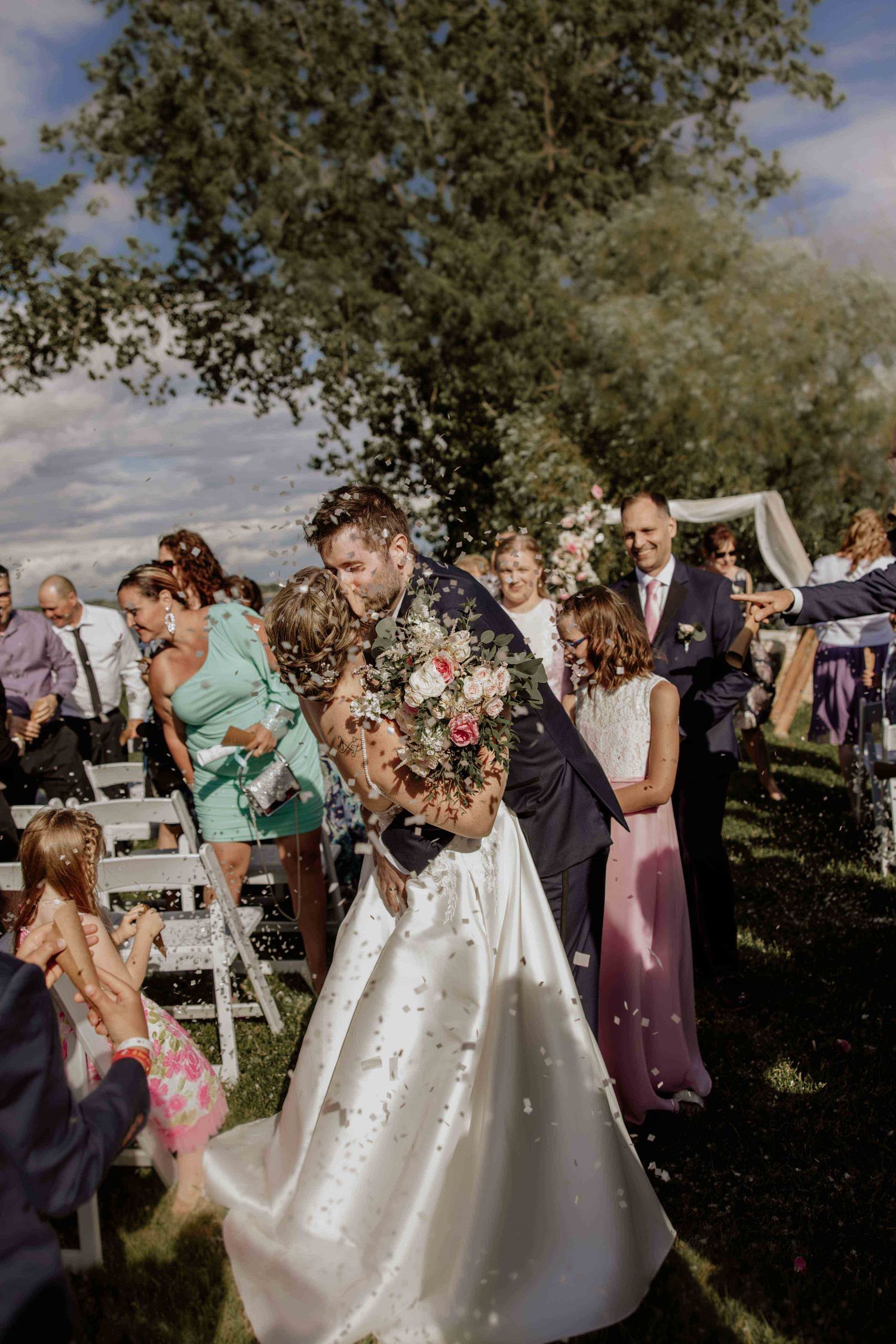Wedding celebration with guests throwing confetti outdoors under blue skies and trees.