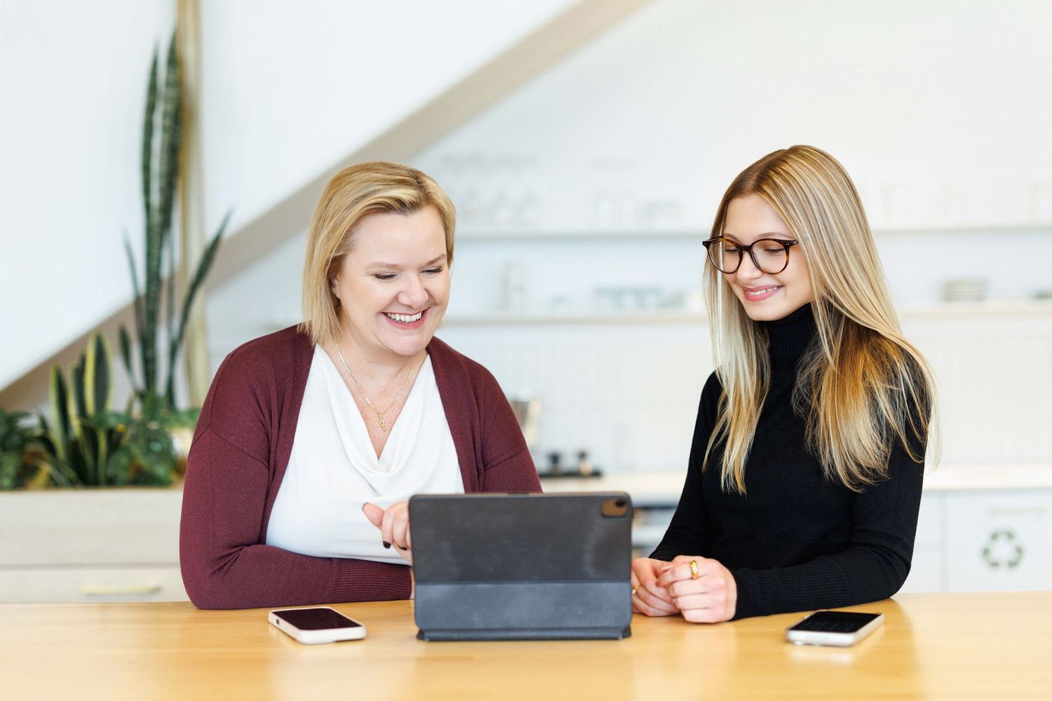 Two professionals in business attire sit at wooden table looking at tablet device during meeting in bright office space.