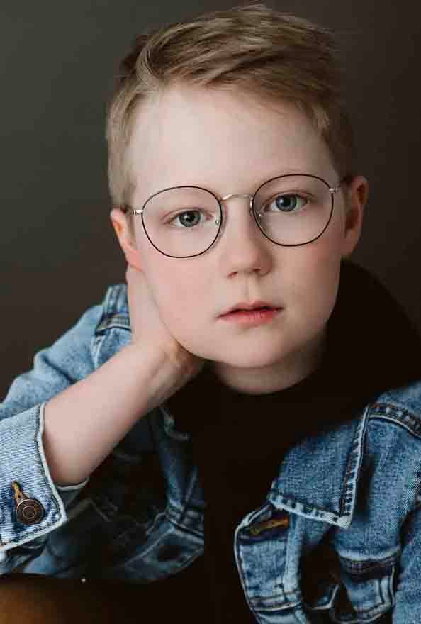 Child wearing glasses and denim jacket posing against dark background in studio portraits.