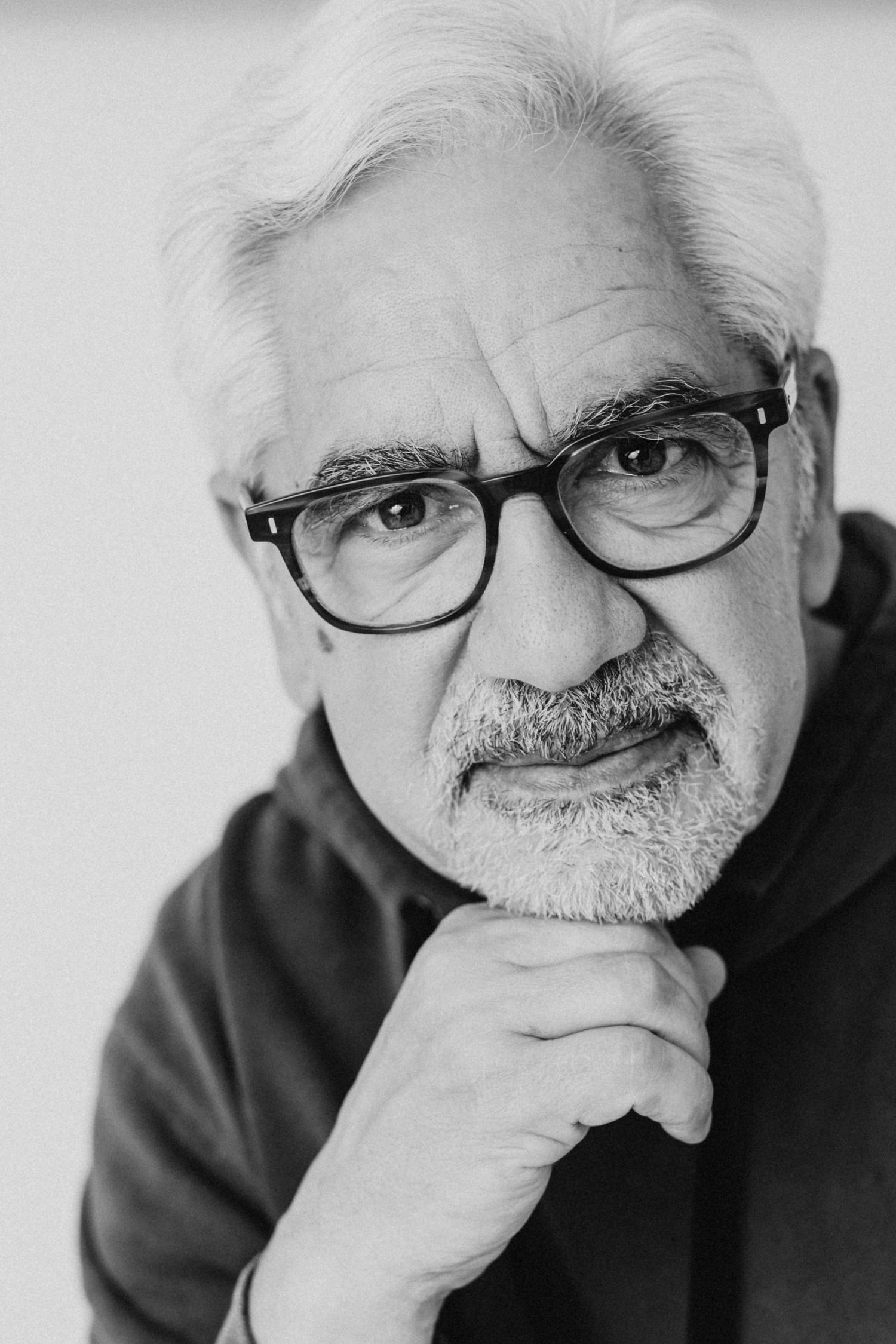 Senior person with glasses and white hair in black top poses thoughtfully against light background.