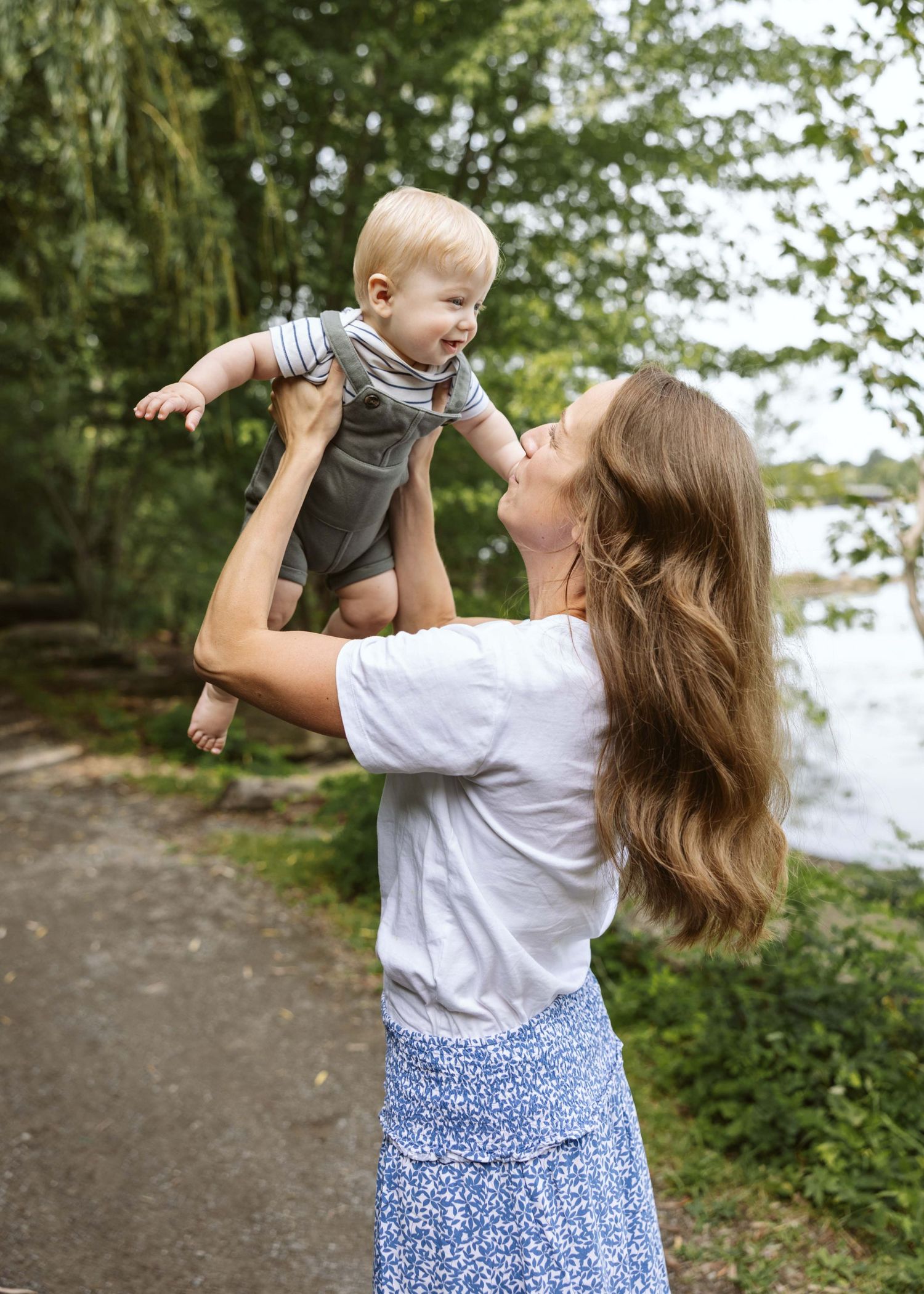 A smiling mother lifts her happy baby boy outdoors near a lake surrounded by lush green trees.