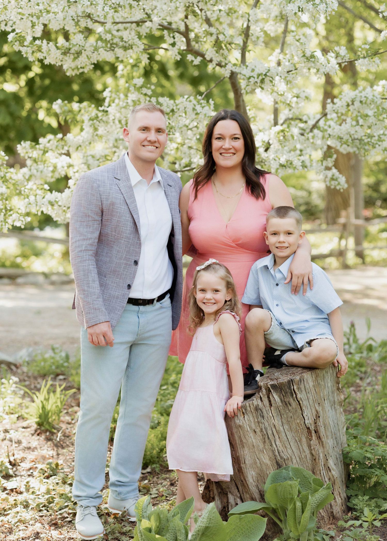 Family of four posing outdoors under blooming white tree, parents with two young children in spring attire.