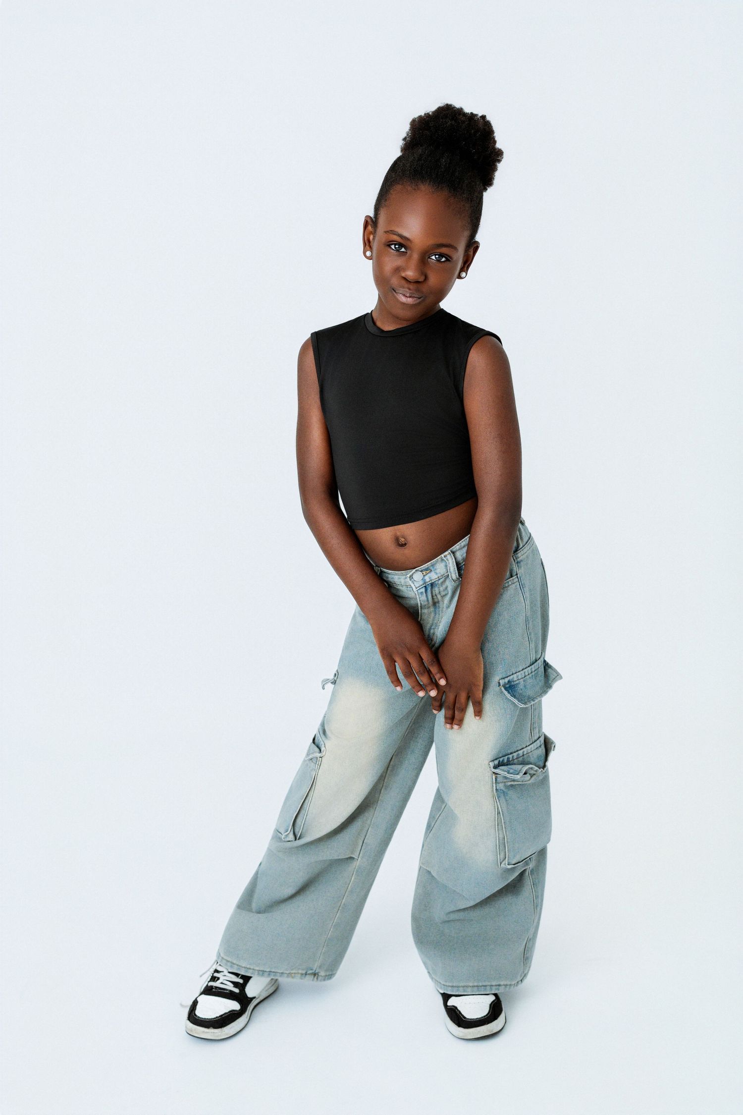 Young girl in black crop top and wide-leg cargo jeans with sneakers posing against white background.