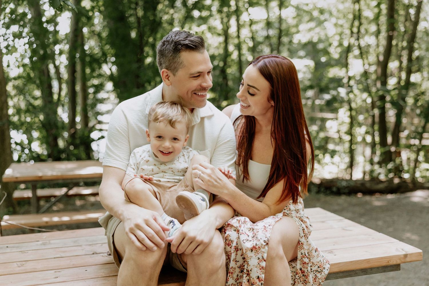 Happy family with a toddler sitting on a wooden bench outdoors, smiling together in a lush green park setting.