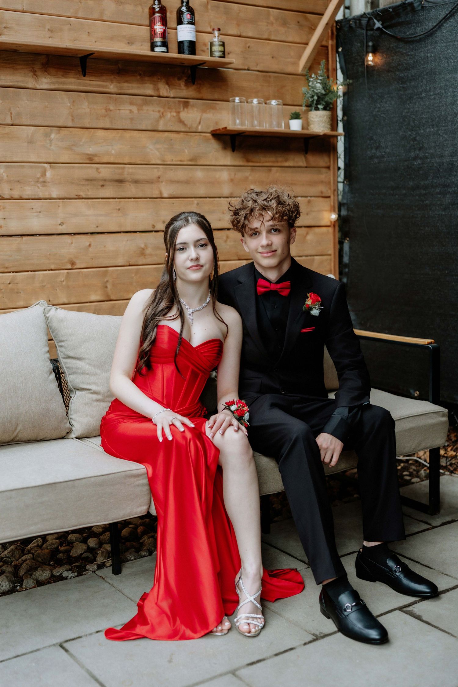 A couple in prom attire, girl in red gown and boy in black tuxedo with red bow tie, seated on a cream sofa.