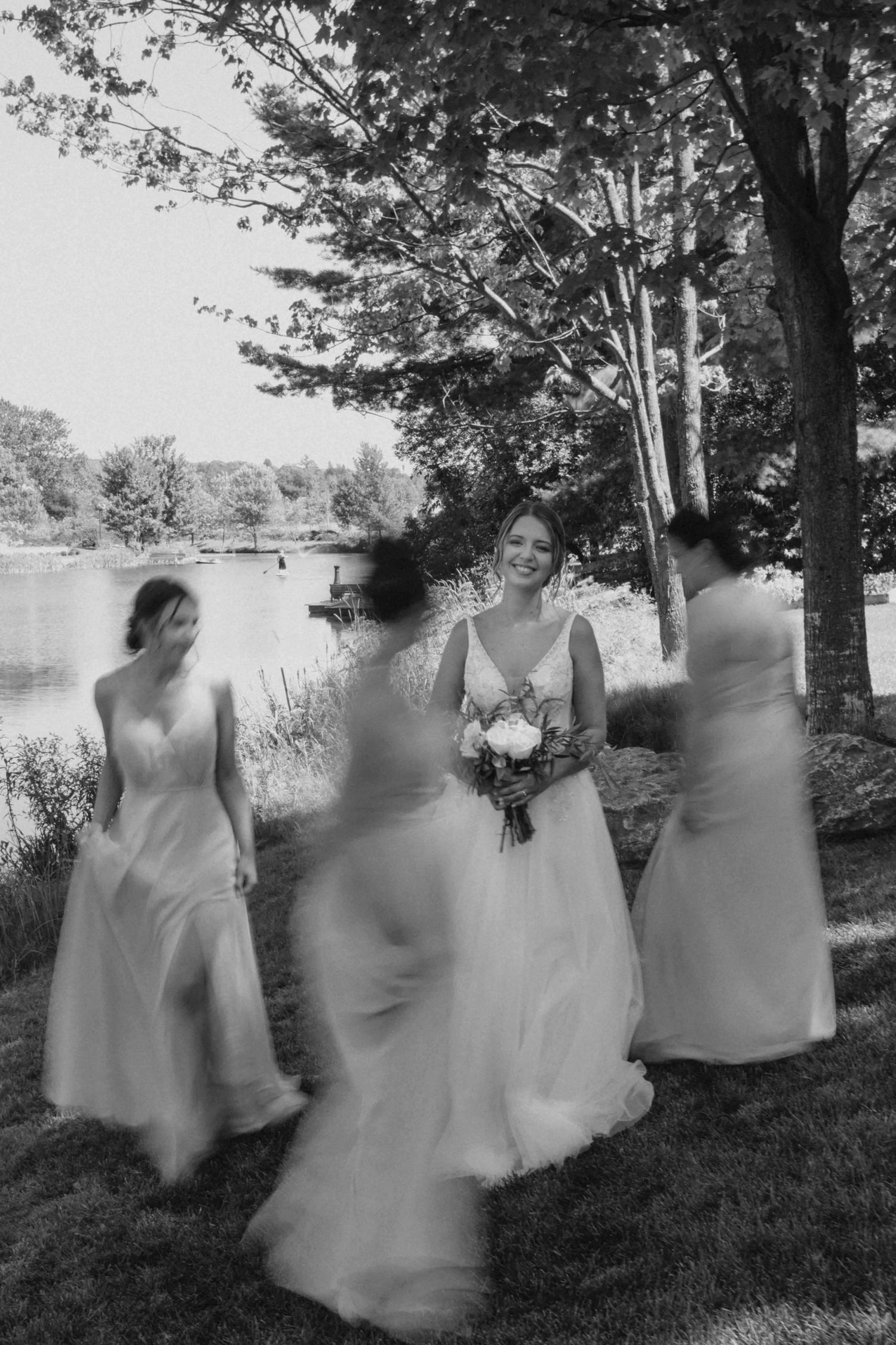 An artistic black and white photo of bridesmaids dancing with the bride on a lakeside lawn.