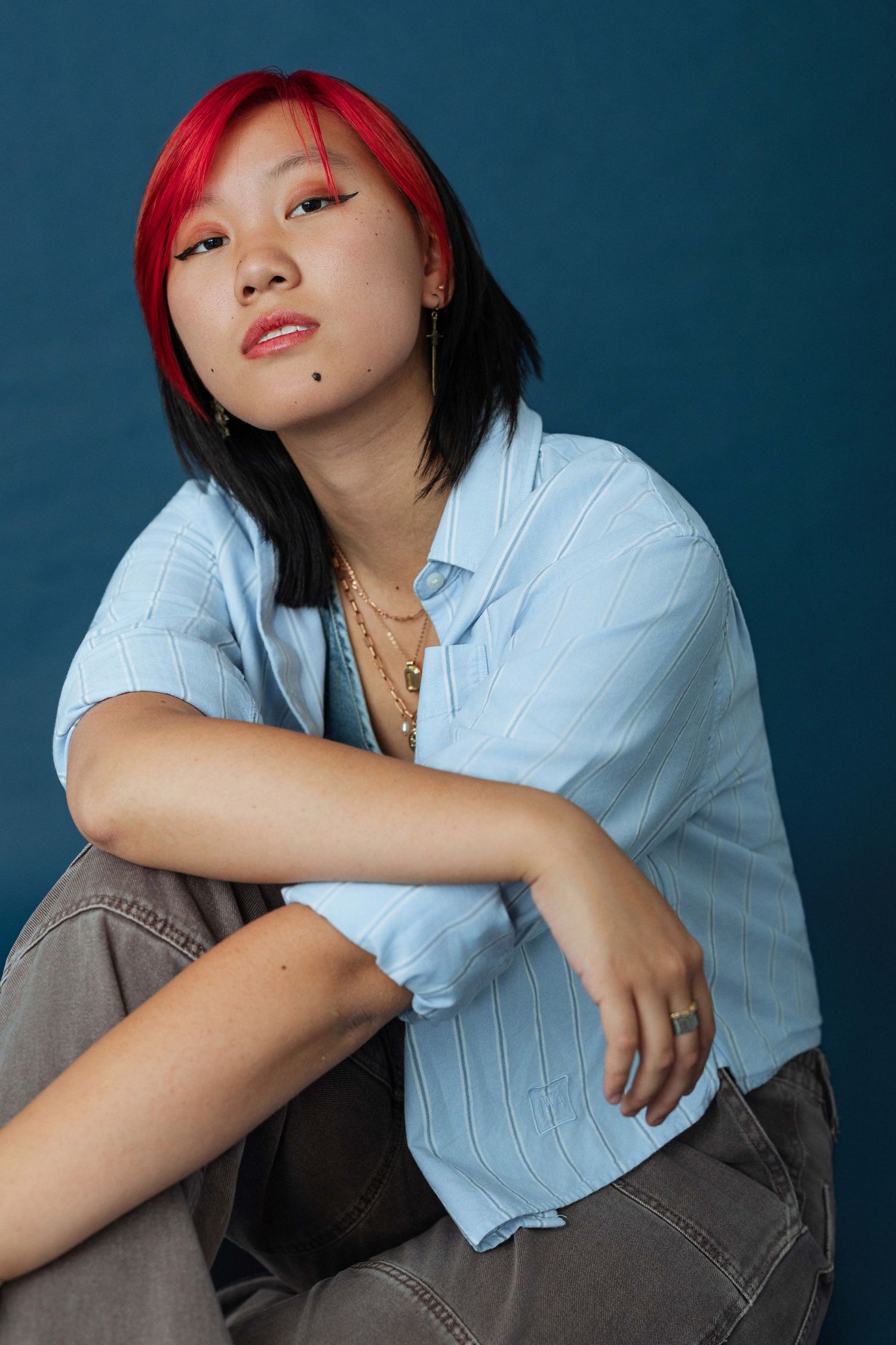 Young woman with red and black hair in blue striped shirt and gray jeans poses on dark blue background.