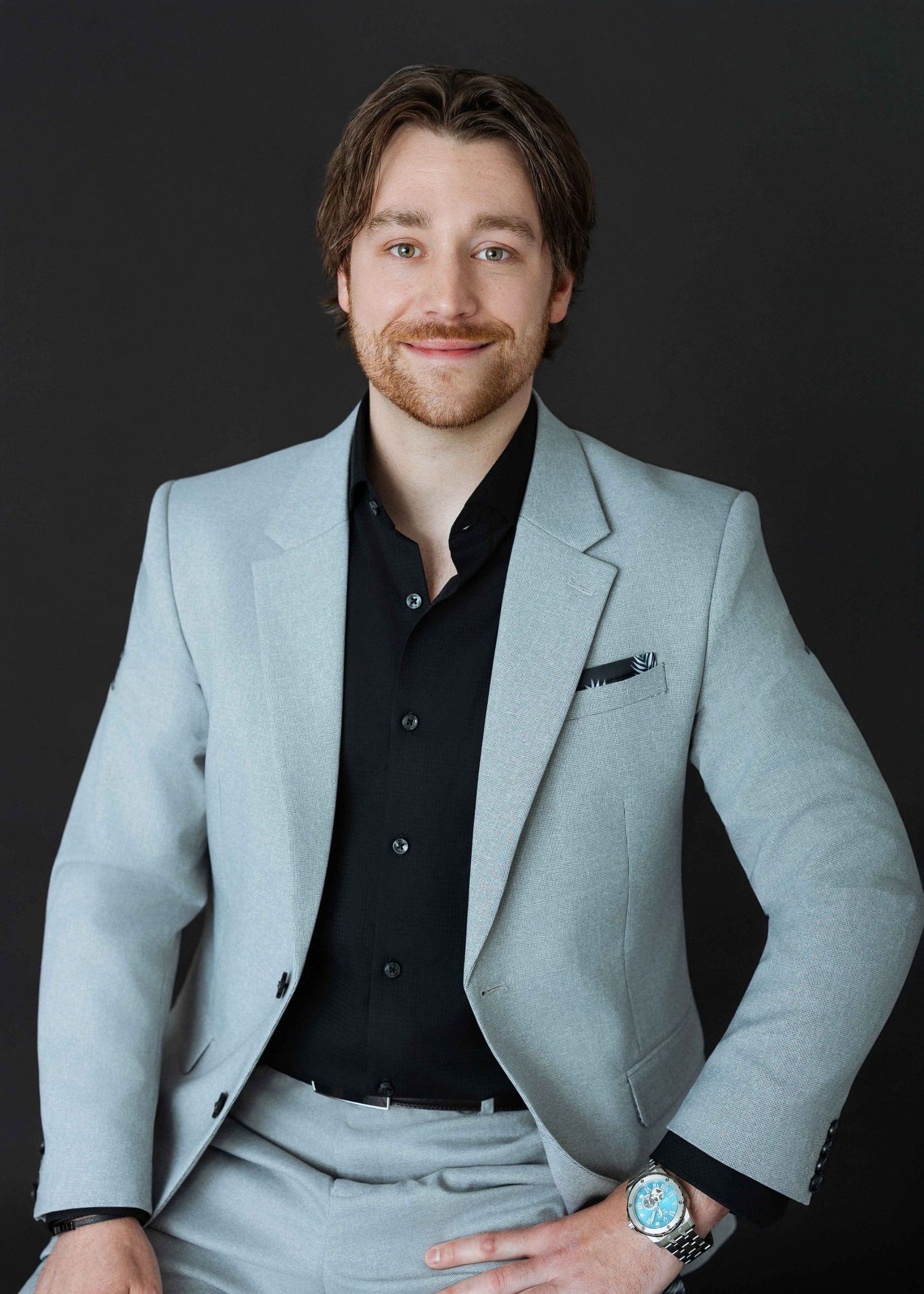 Professional man in light gray suit and black shirt poses confidently against dark background for business portrait.