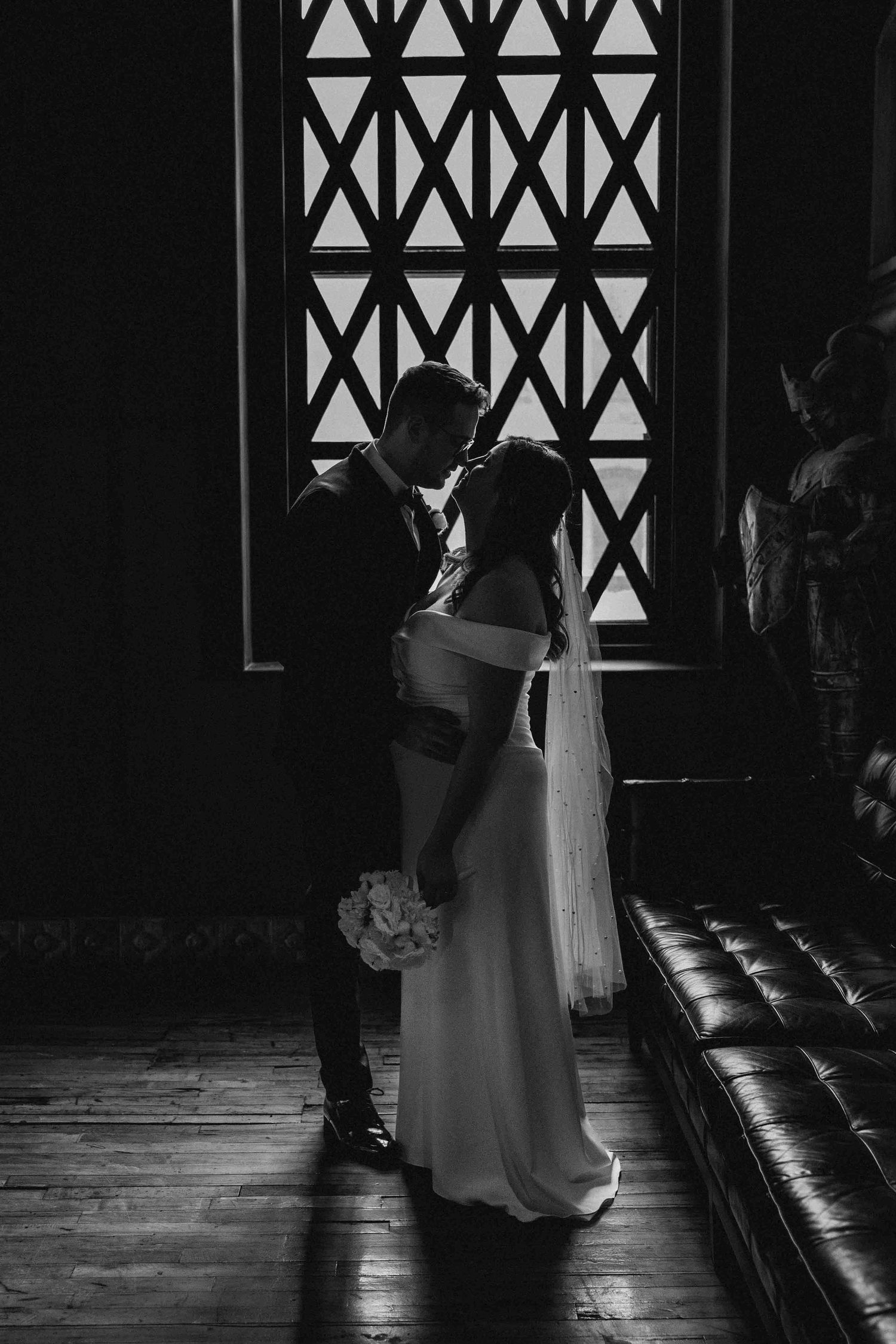 Black and white wedding photo showing couple's silhouette against window with geometric pattern in dramatic lighting.