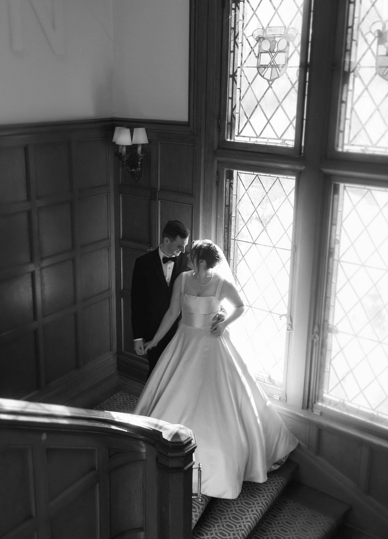 A couple in wedding attire stands by a large leaded glass window in a wood-paneled stairwell, captured in black and white.