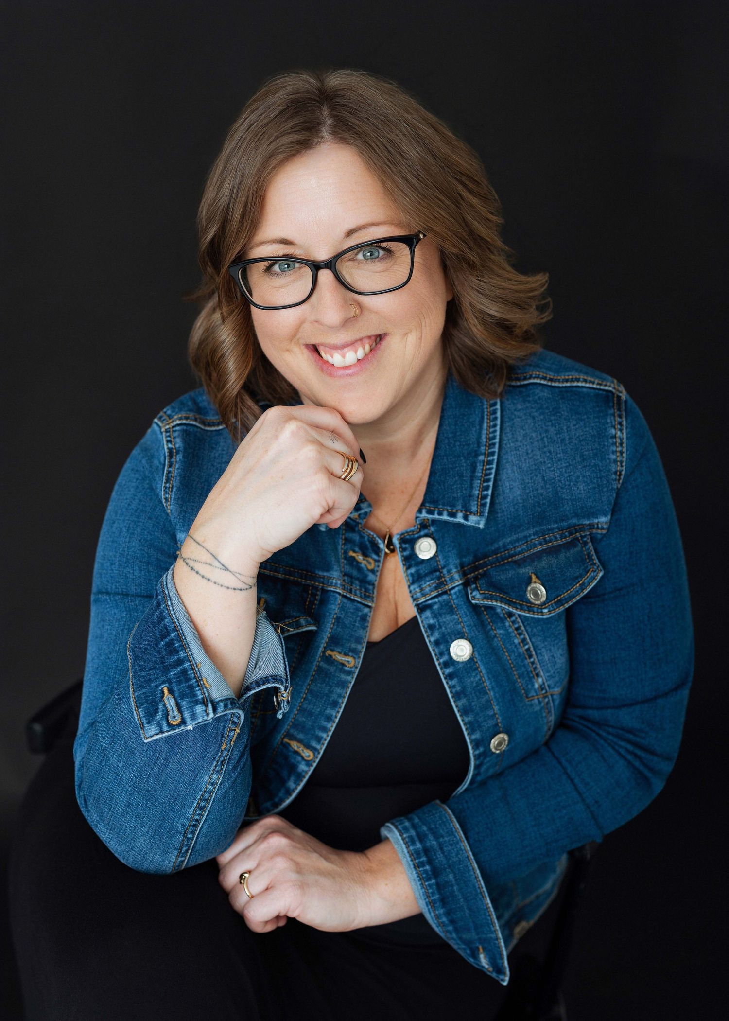 Smiling woman with glasses wearing a denim jacket poses confidently against a dark background.