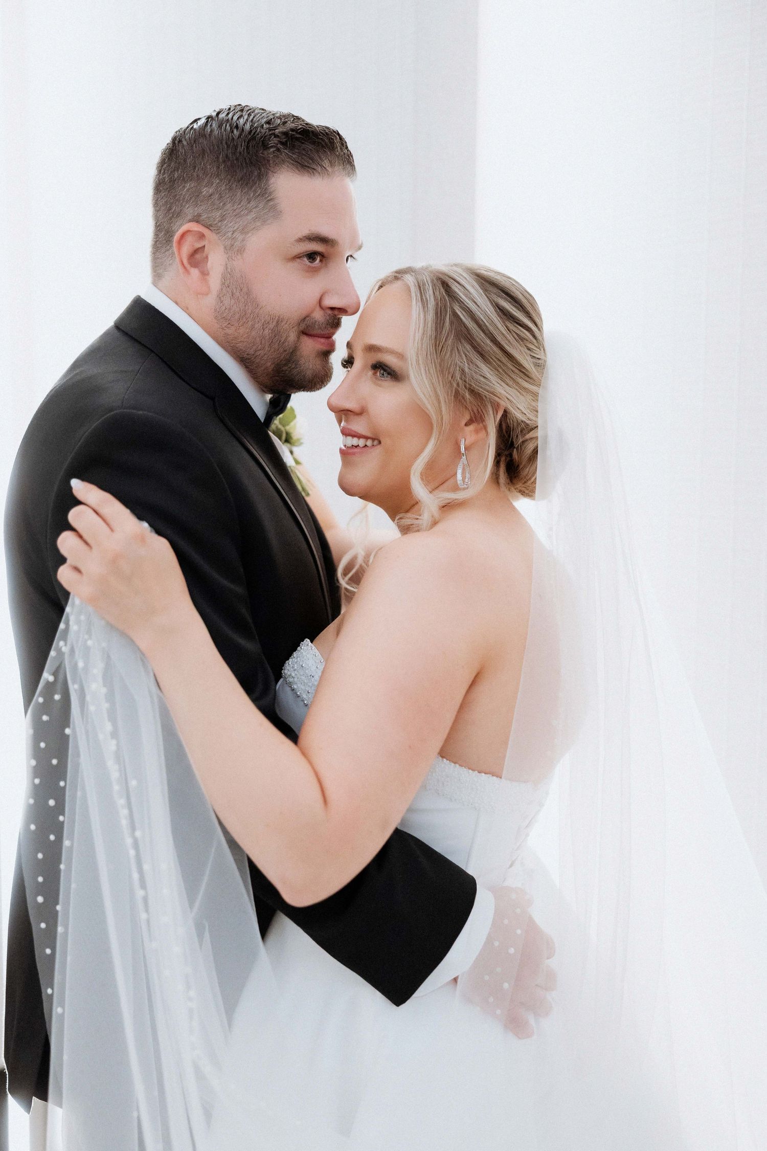 A romantic wedding portrait of a couple embracing against a white wall with the bride wearing a flowing veil.