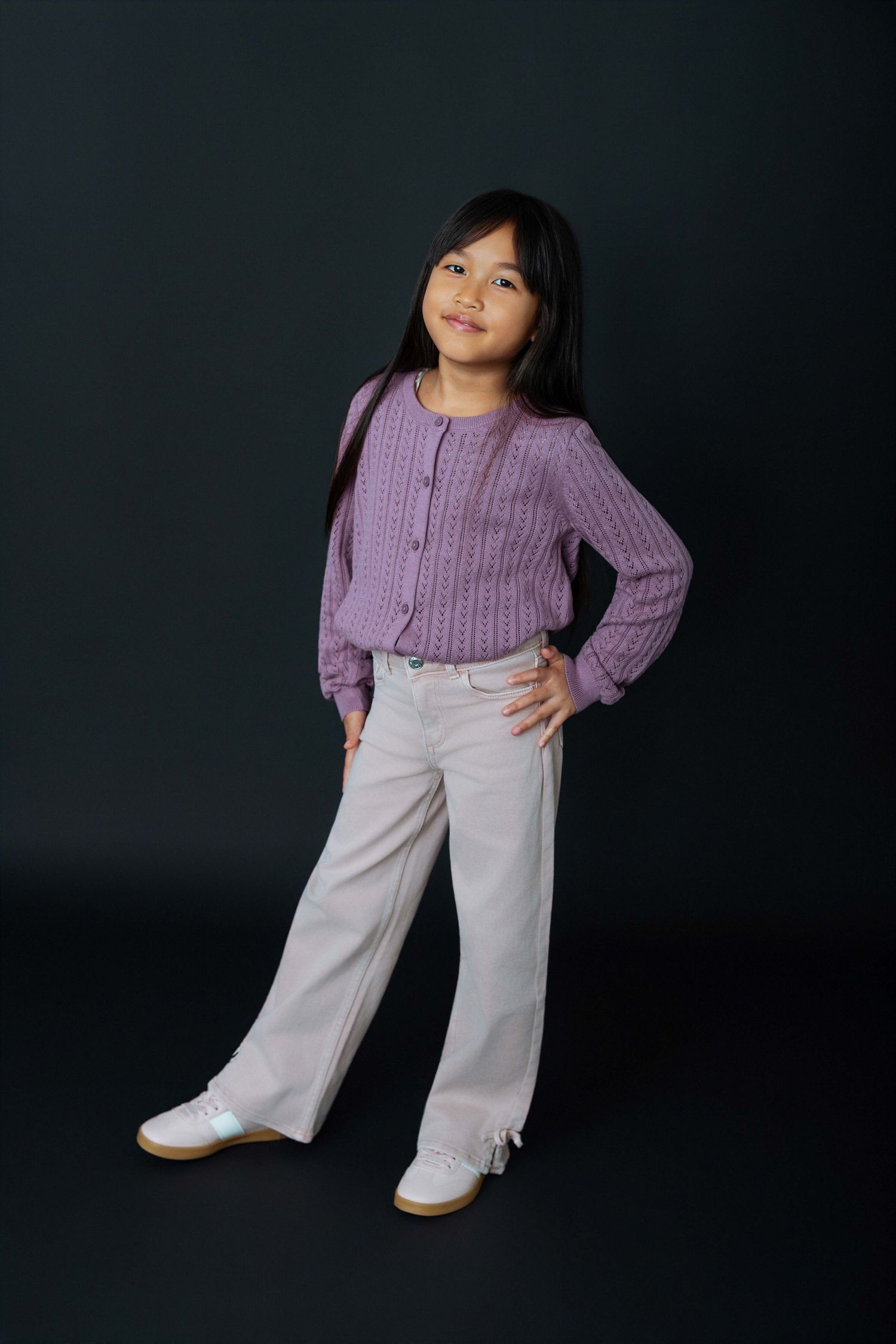 Young girl in purple knit cardigan and wide-leg cream pants poses confidently against dark background.