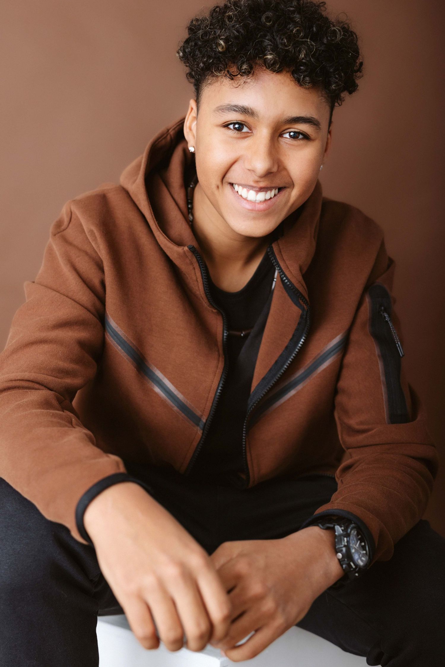 Professional studio portrait of a person in a brown jacket and black shirt smiling warmly against dark background.