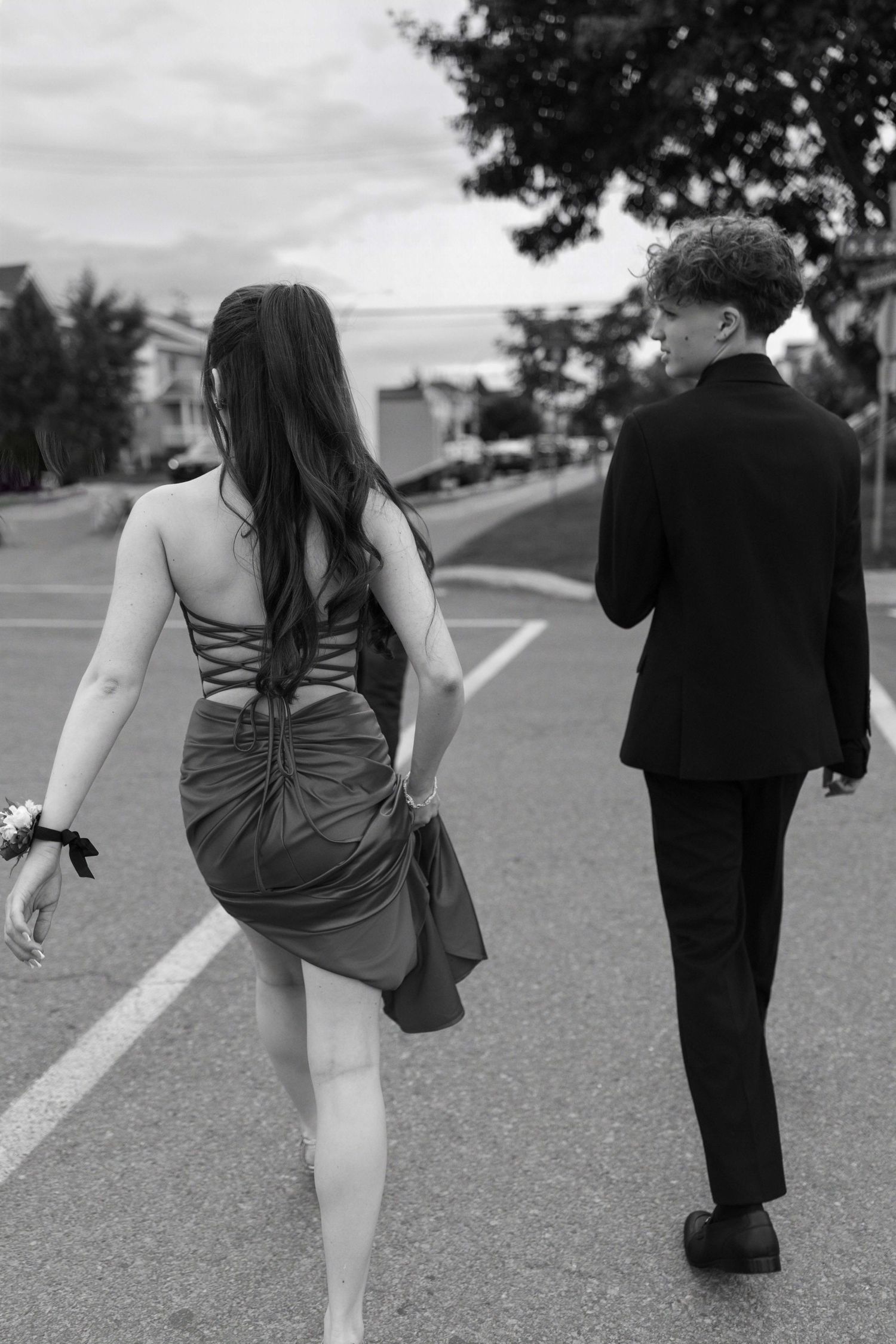 Black and white photo of a couple in formal attire walking on a road, woman in backless dress, man in suit.