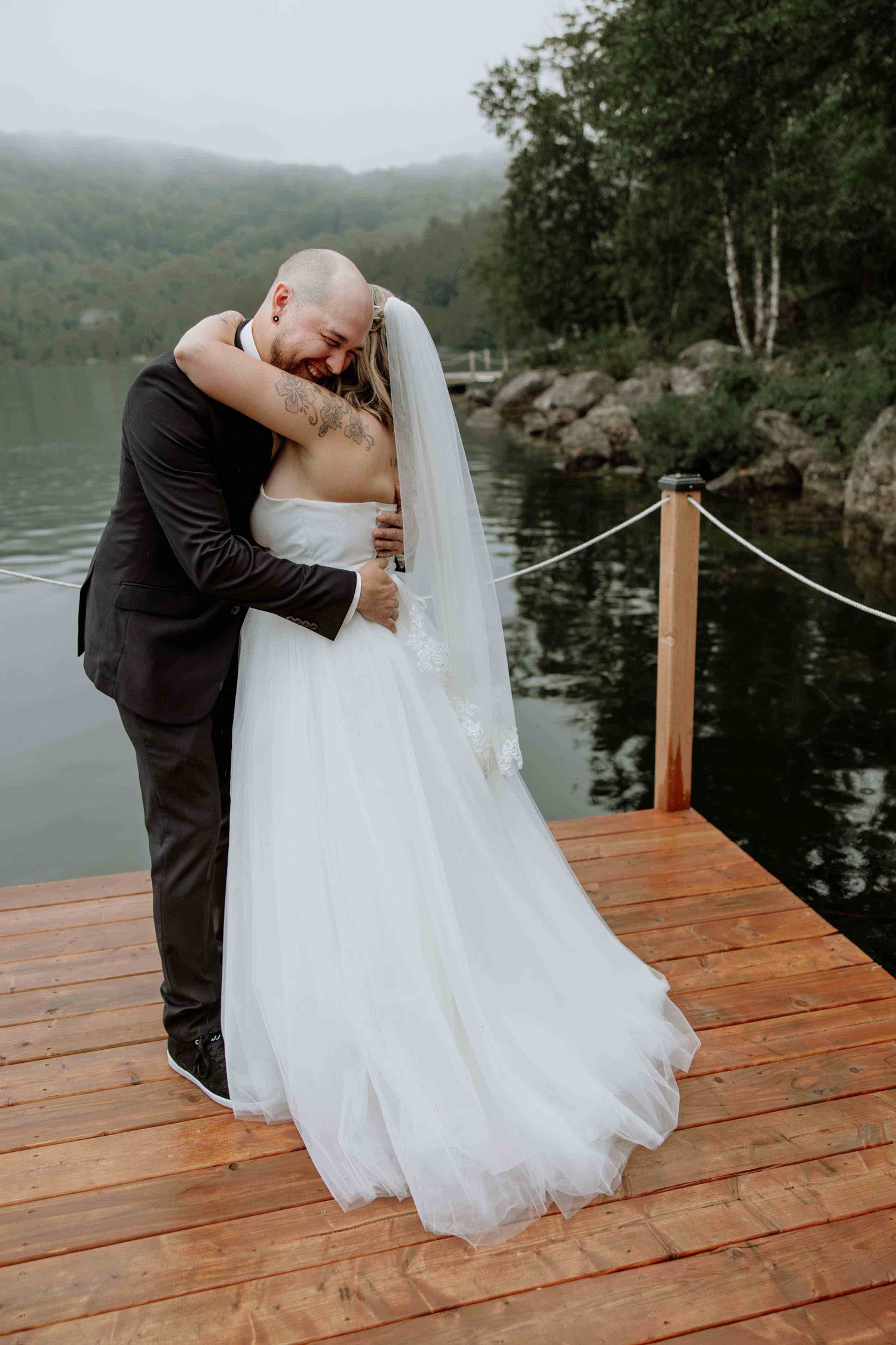 Romantic wedding couple shares intimate moment on wooden dock overlooking misty mountain lake.